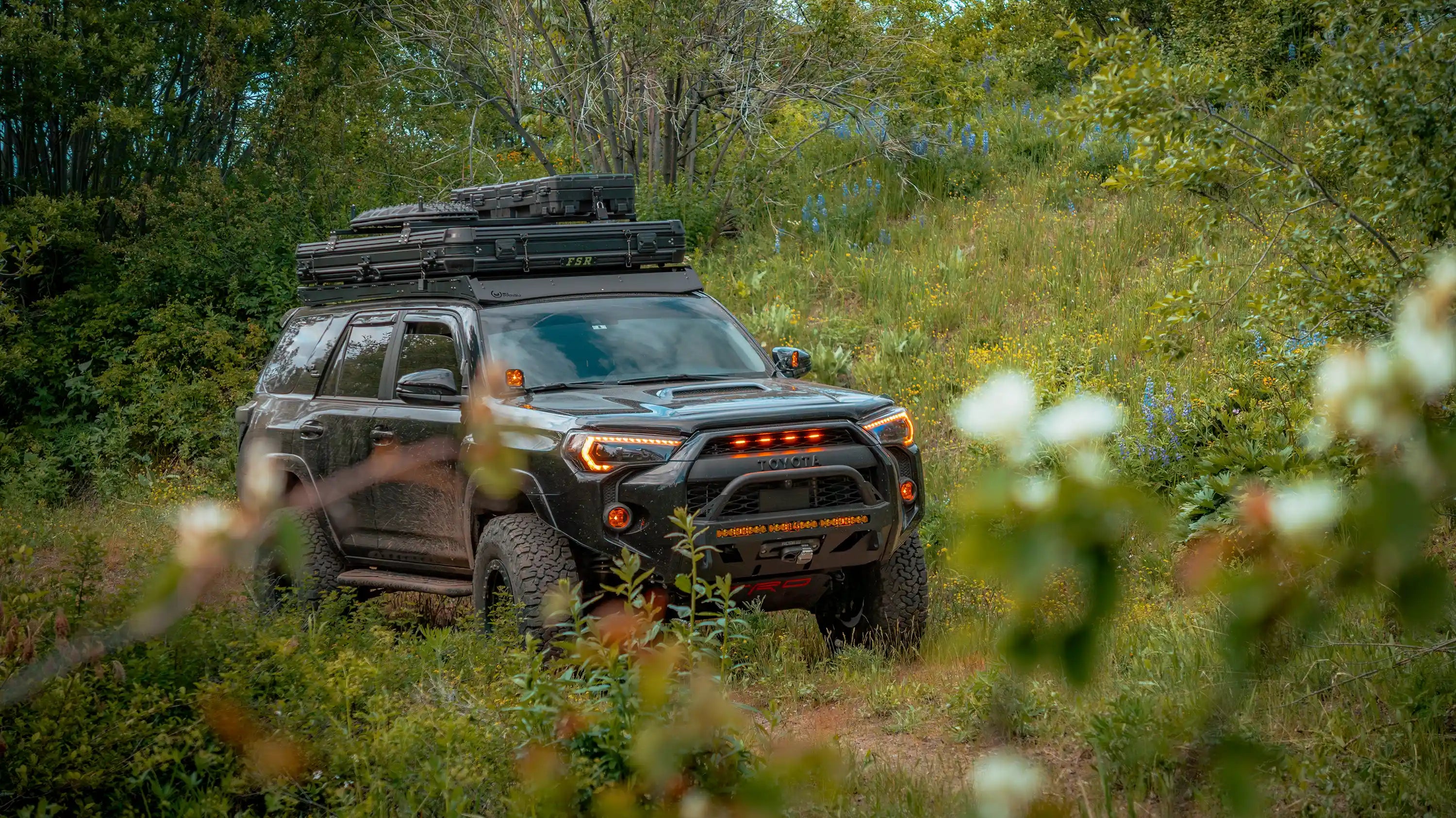 Toyota 4Runner driving off road through a forested area with a FSR Rooftop Tent mounted on the roof rack and traction boards installed on top of the roof top tent