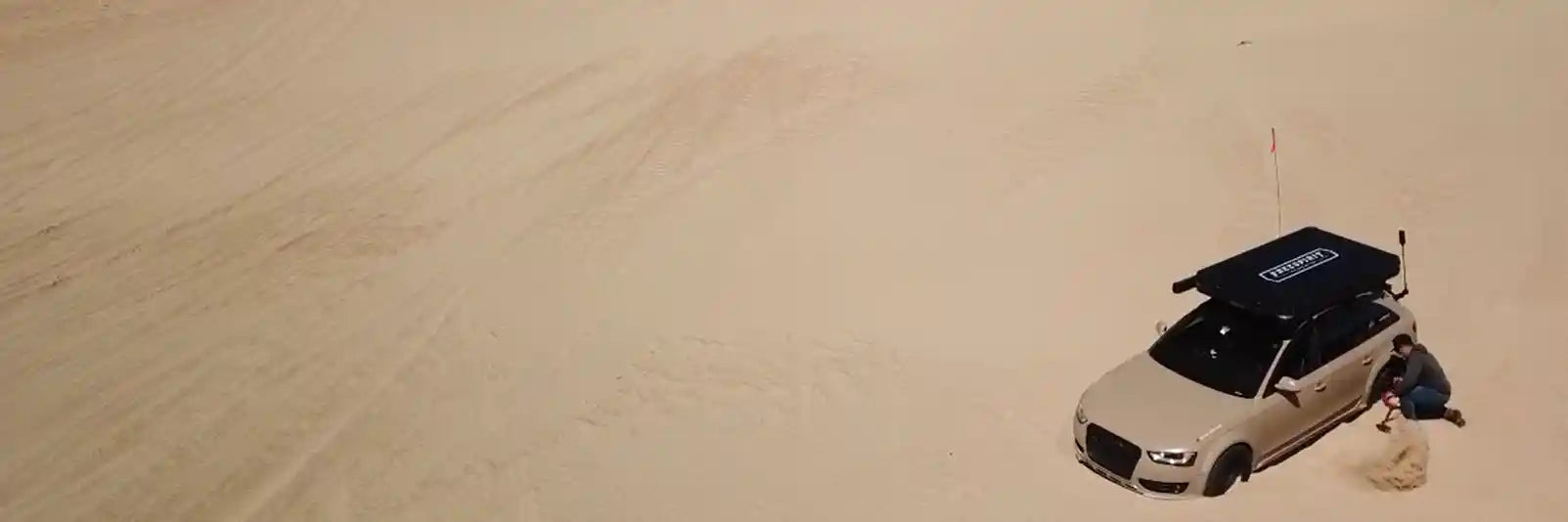 Car with a roof rack mounted FSR rooftop tent stuck in the sand and being dug out by a person in a desert landscape