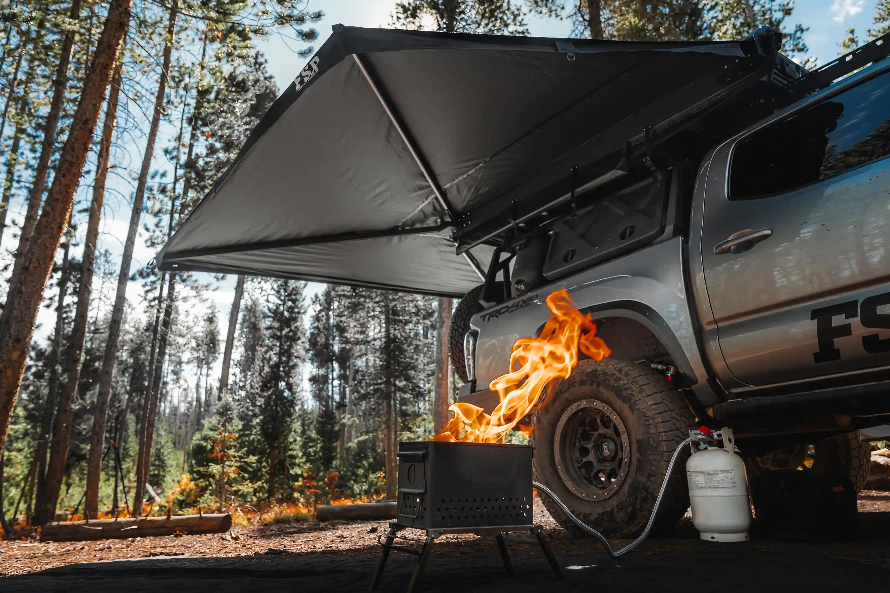 Freespirit Recreation 270 Awning installed on a Toyota Tacoma deployed while camping in a forested setting with a propane firepit visible in the foreground