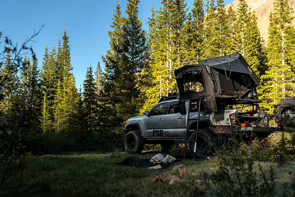 Silver Toyota Tacoma with an FSR Kali roof top tent installed on the bed rack and setup for camping in a forested area with a mountain visible in the background.