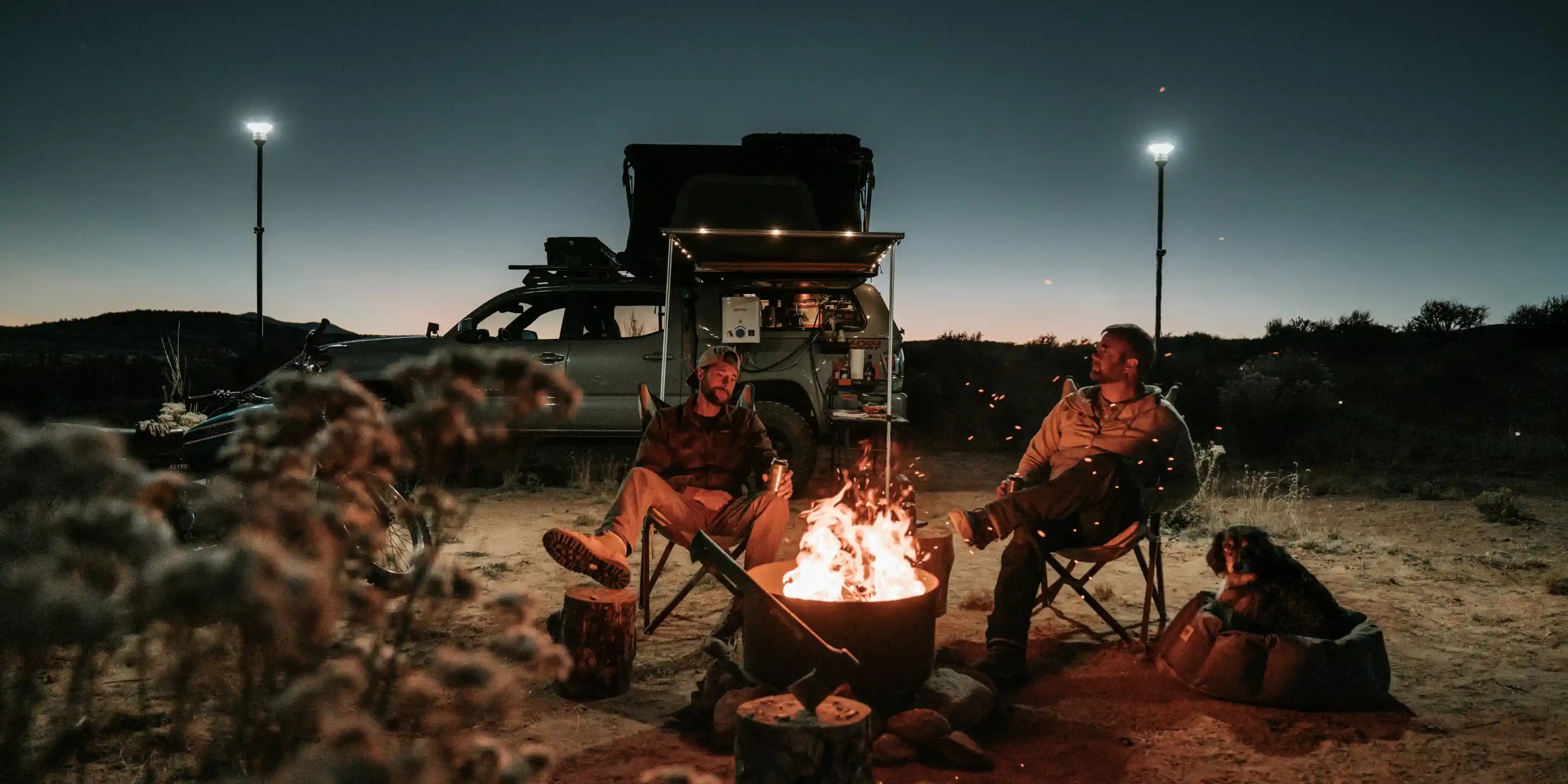 Two people and a dog sitting around a campfire at night with a Toyota Tacoma in the background equipped with a Freespirit Recreation rooftop tent and awning