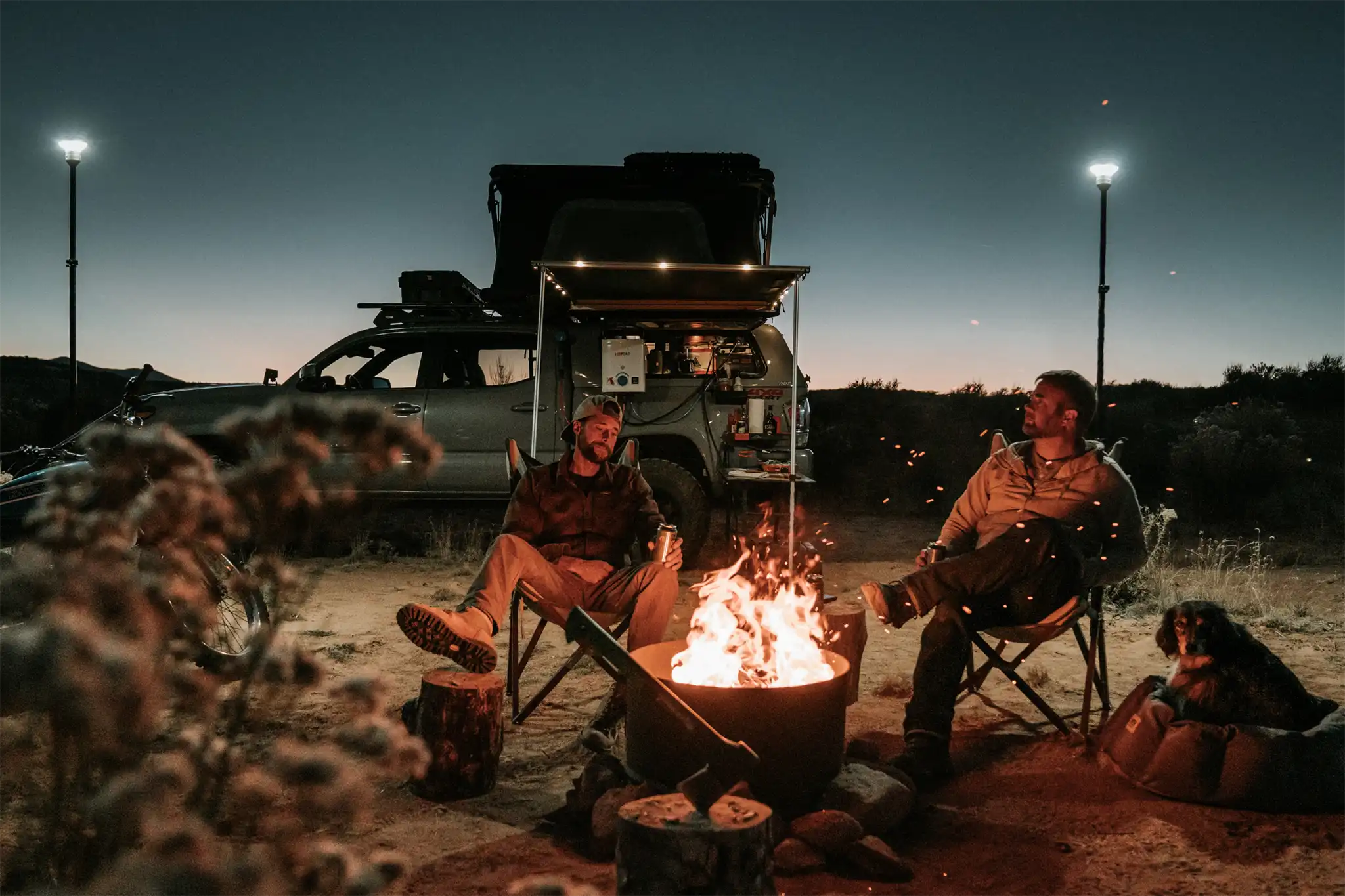 Two men sitting around a campfire at night with a Toyota Tacoma fitted with an FSR Rooftop Tent in the background.