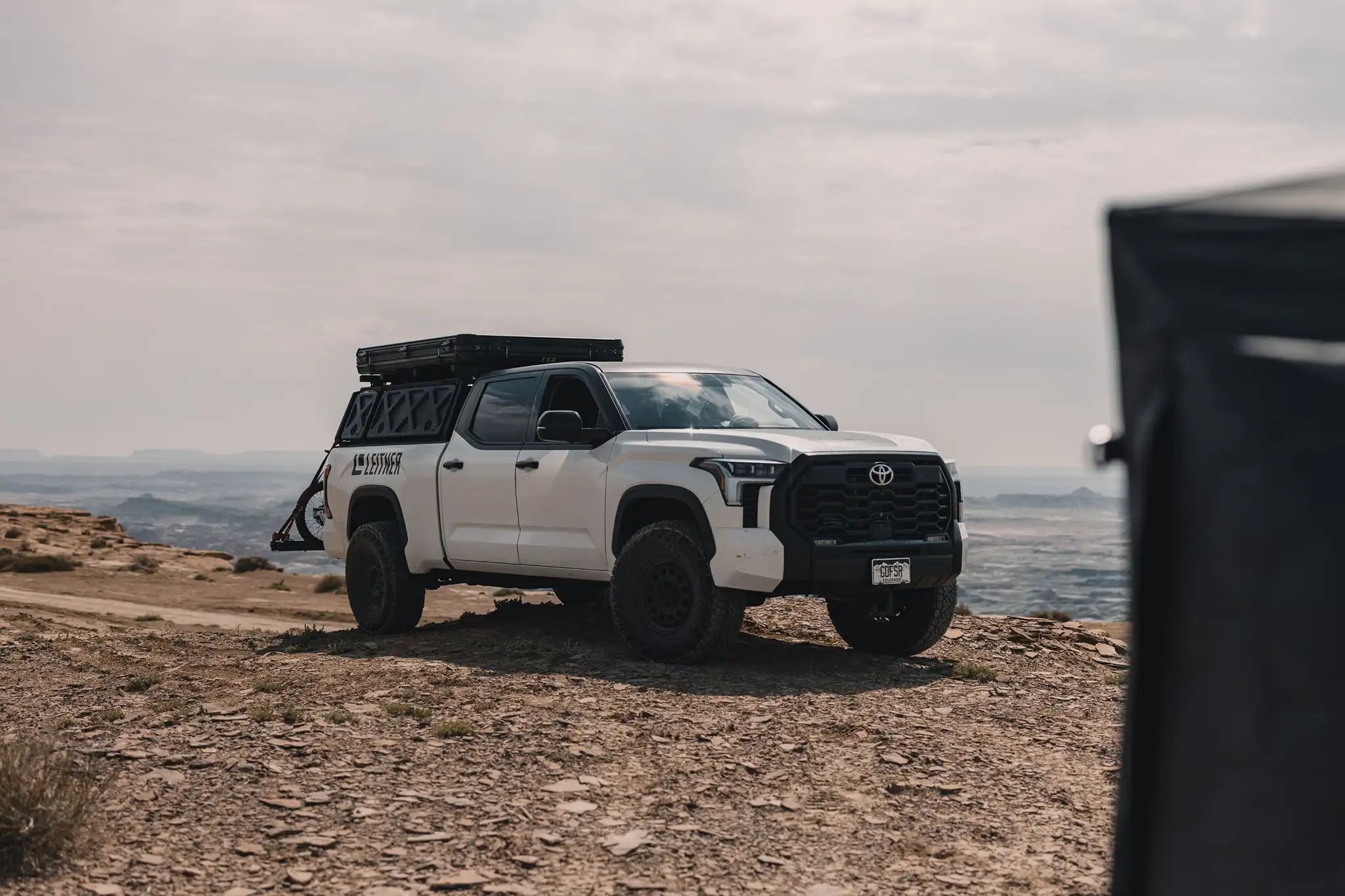 White Toyota Tundra with a bed rack installed and a Free Spirit Recreation Rooftop Tent mounted for camping on a rocky campsite overlooking desert terrain