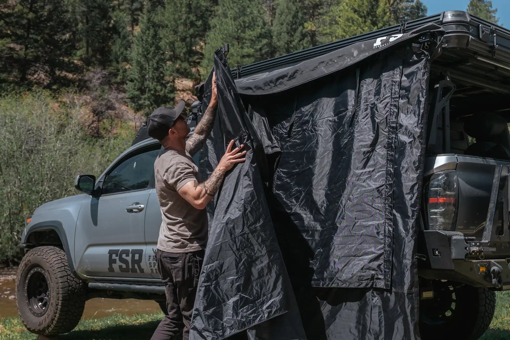 Person setting up the FSR Double Shower Awning & Privacy Tent installed on a Freespirit Recreation Roof Top Tent while camping with a river visible in the background