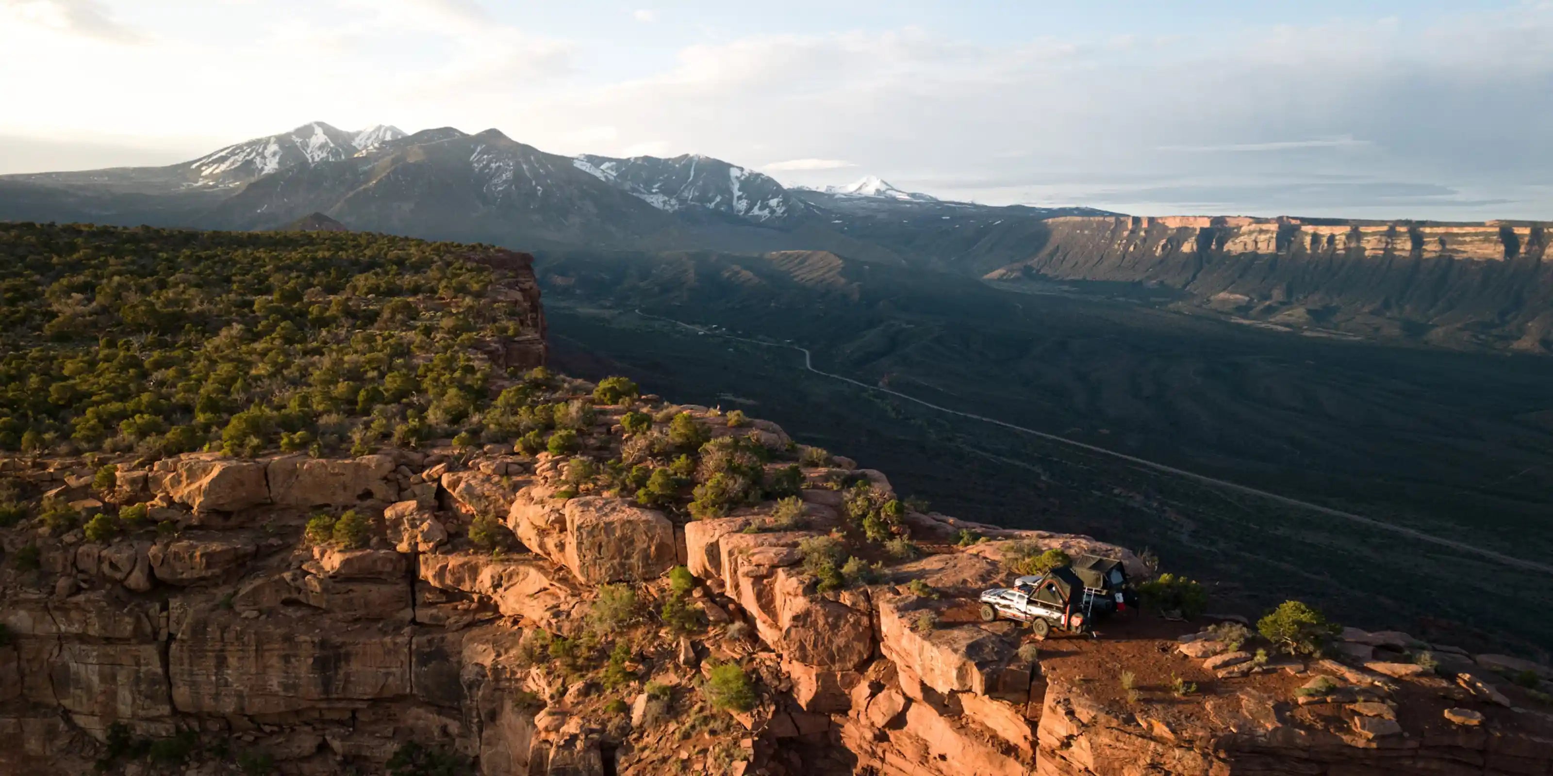 Scenic view of a canyon with red rock formations and snow-capped mountains in the distance and two trucks equipped with FSR Rooftop Tents and Awnings setup camping on a red rock ridge in the foreground.