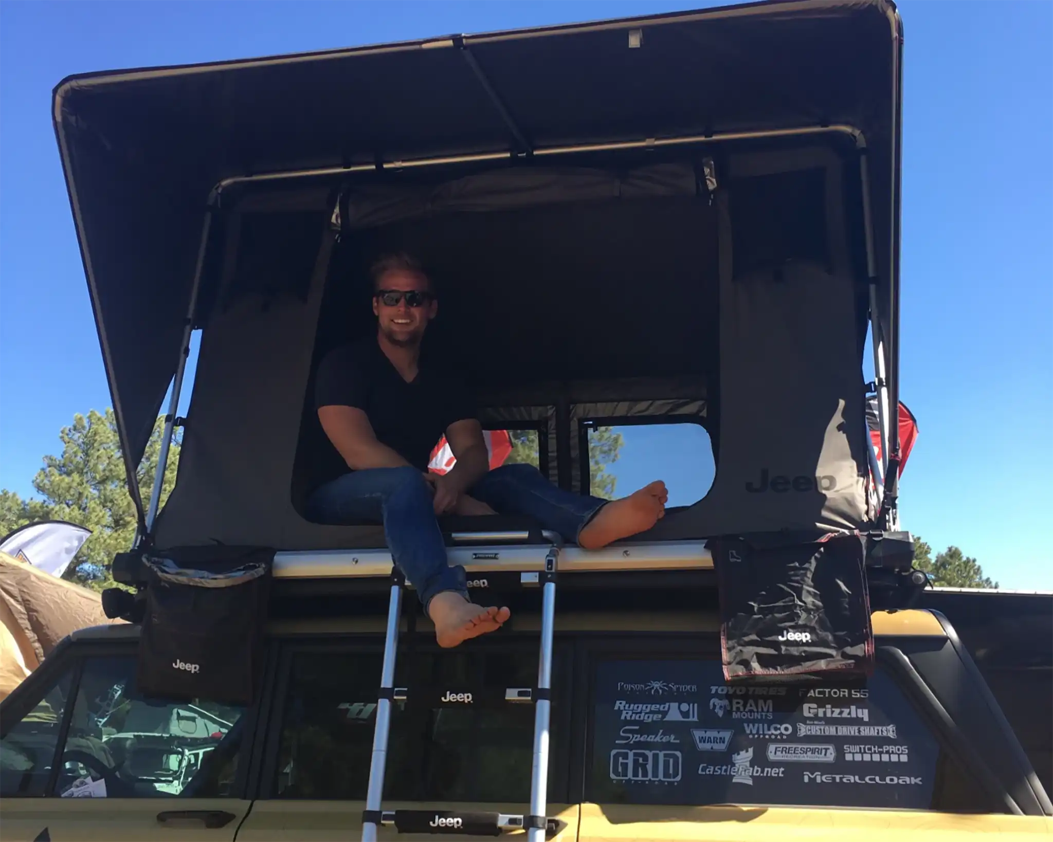 Person sitting inside a Free Spirit Recreation rooftop tent on a vehicle roof rack at an Overland Expo with a clear blue sky in the background in 2015