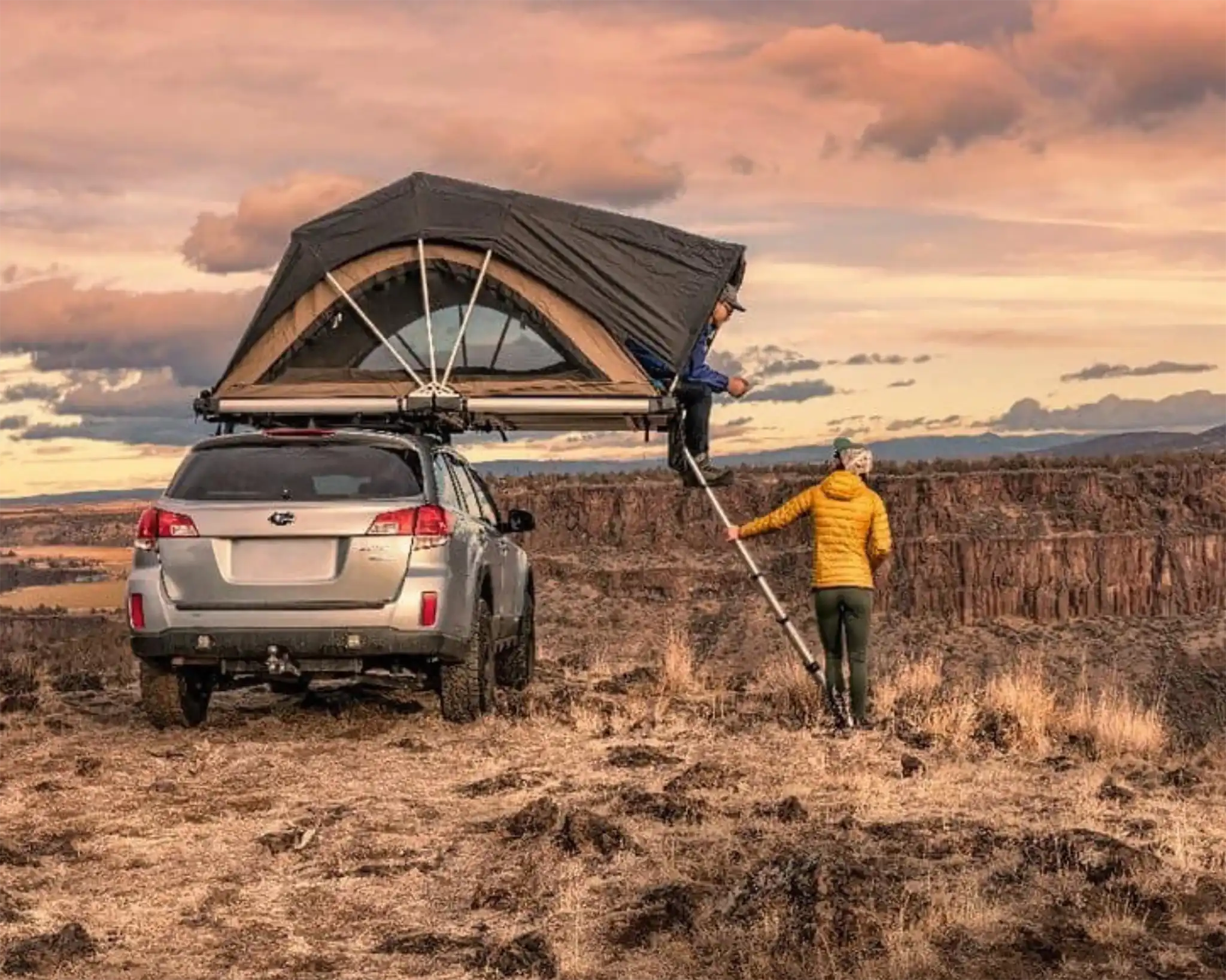 Two people setting up an FSR High Country rooftop tent mounted on a Subaru Forrester in a desert landscape during sunset in 2018
