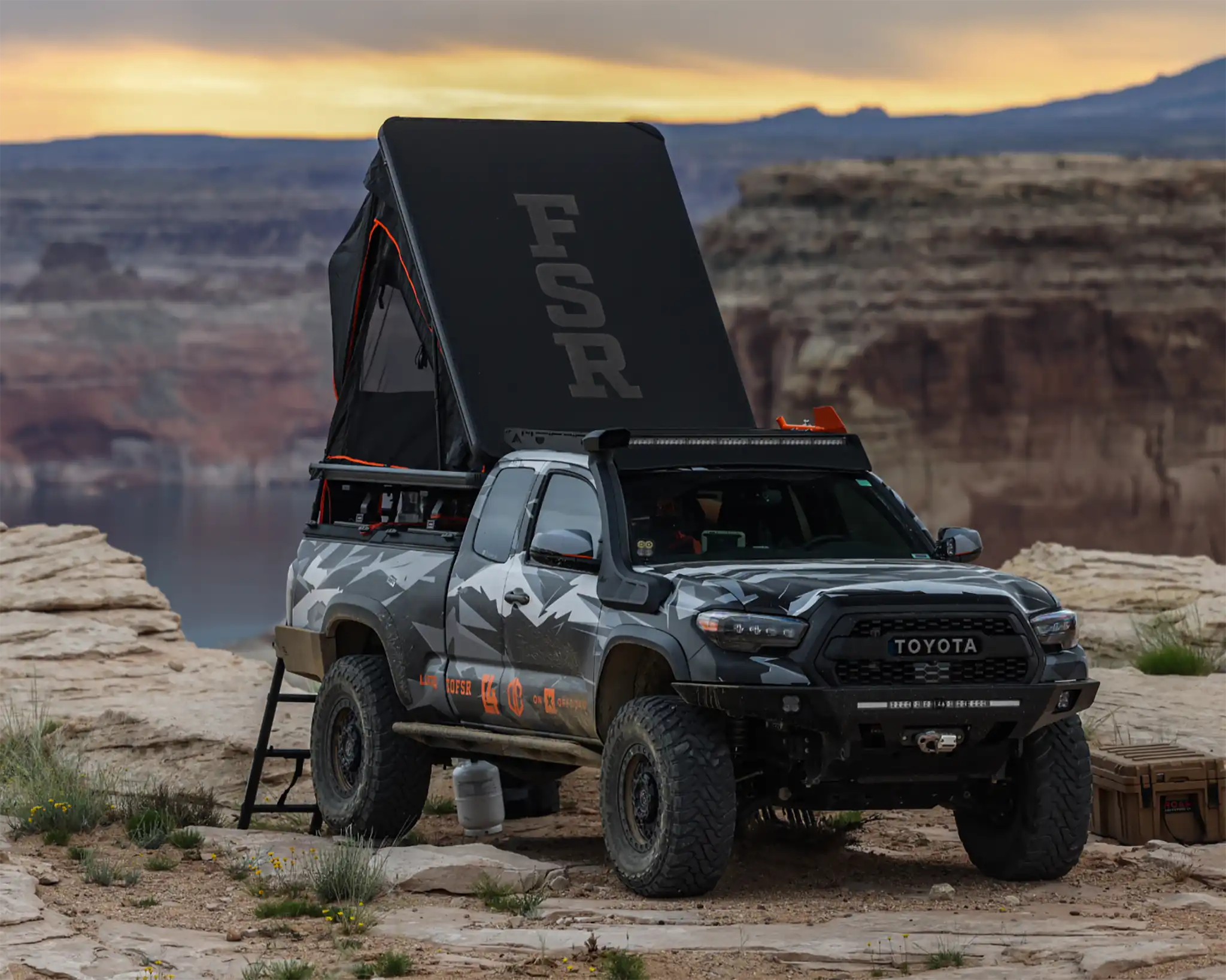 A 2023 FSR Aspen Rooftop Tent mounted on a Toyota Tacoma overland truck setup for camping on a red rock ridge with a body of water and mountainous rock formations in the background