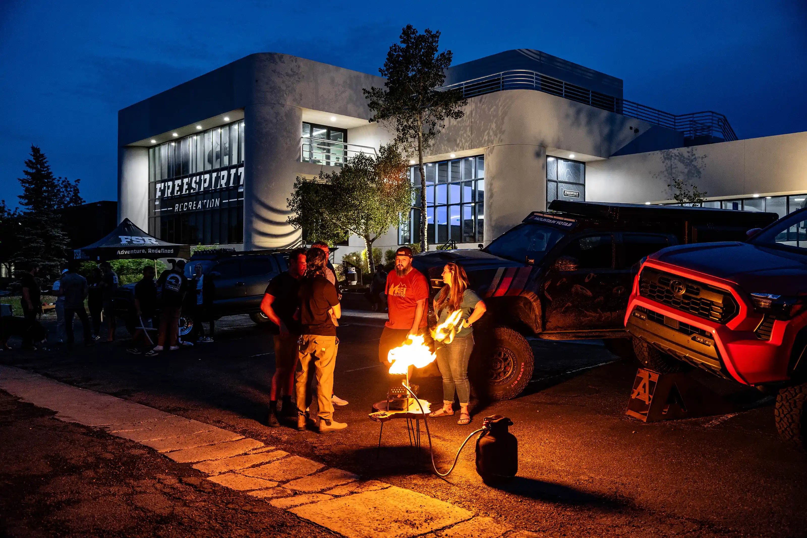 Group of people gathered around a propane fire pit in a parking lot outside the old Free Spirit Recreation headquarters building at night.