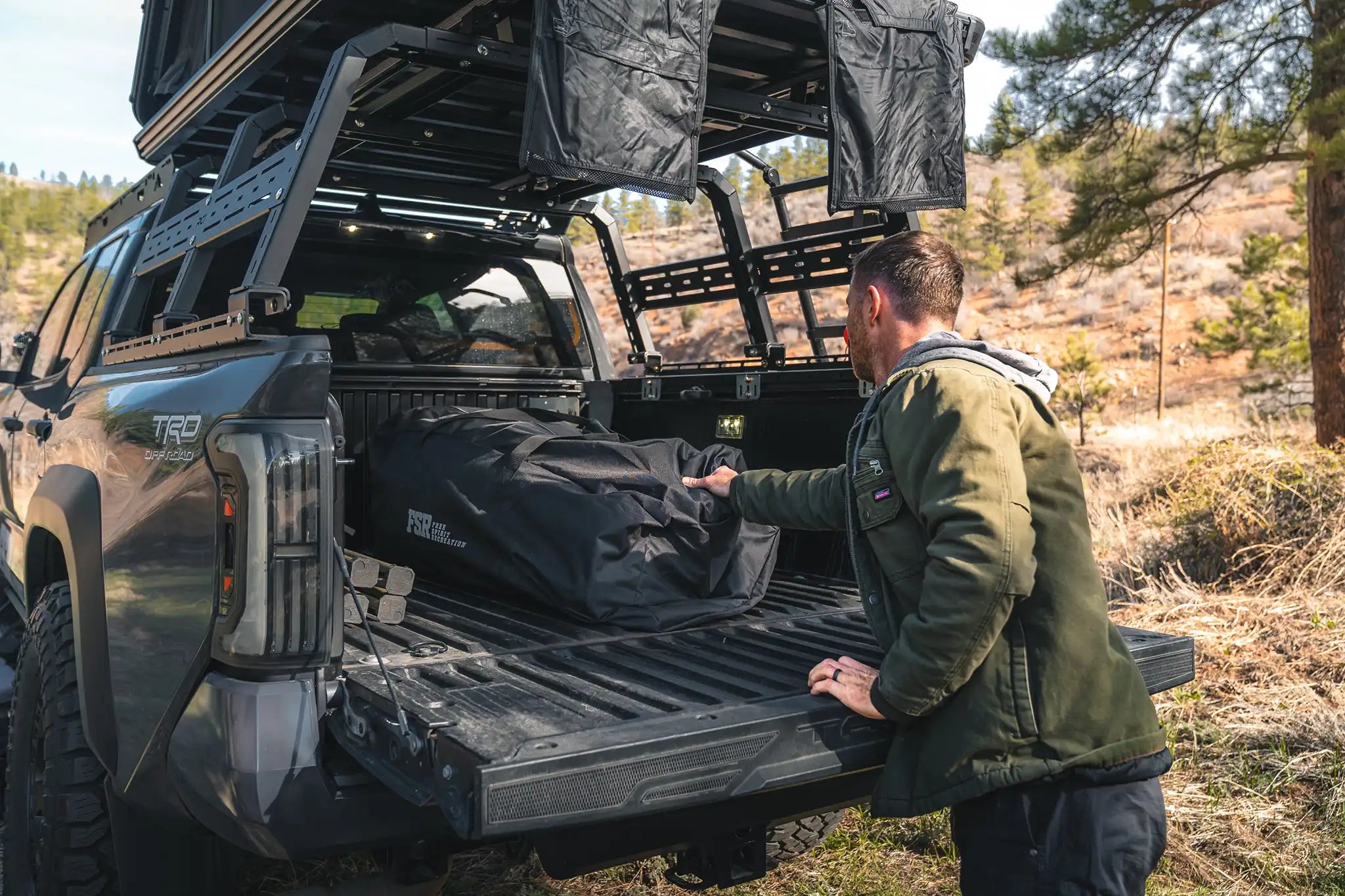 Man removing the FSR Hub 4 Double Tent from Freespirit Recreation, packed in its storage bag, from the back of a truck bed in a wooded campsite.