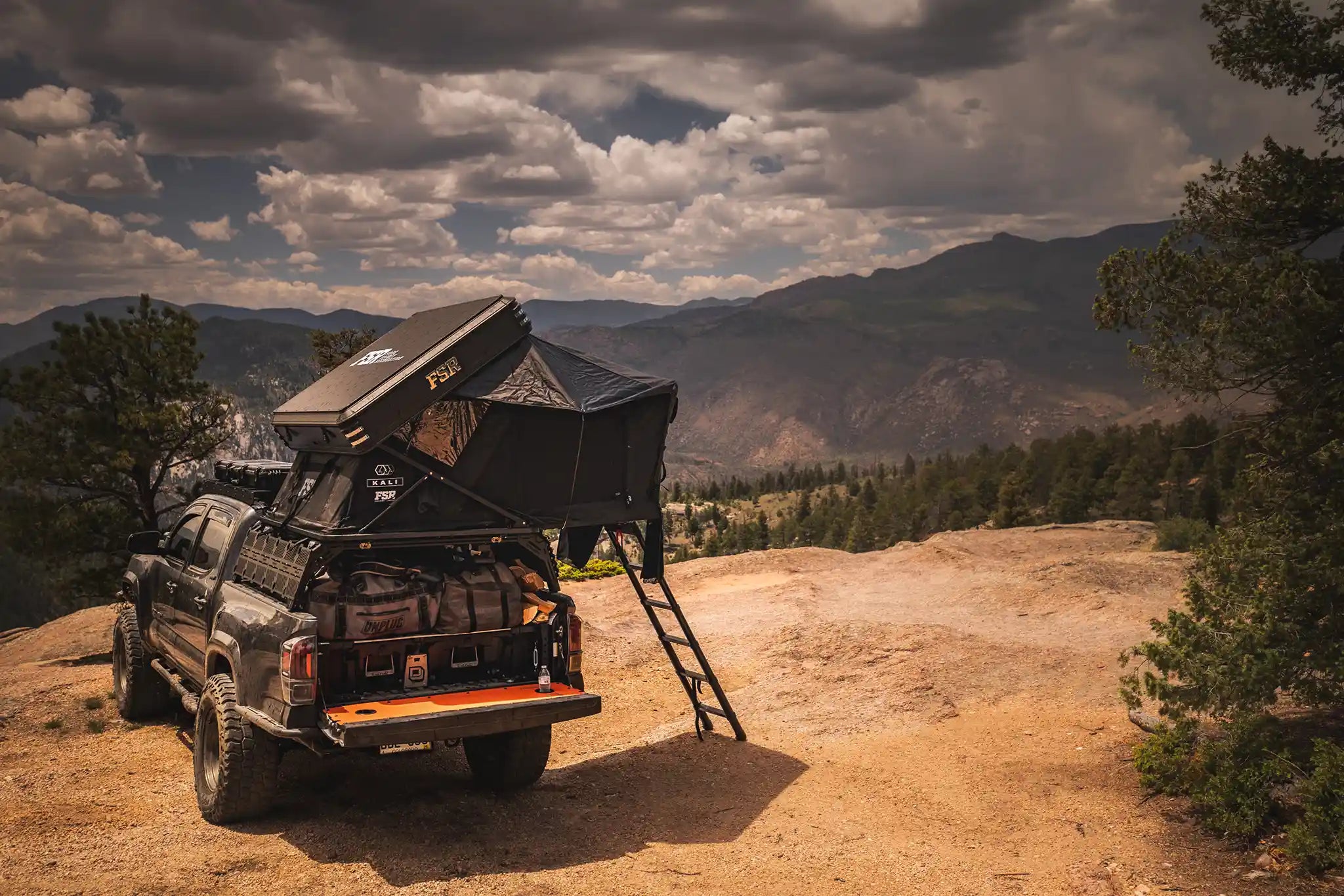 Rear-facing image of a Toyota Tacoma overlanding setup with an FSR Kali rooftop tent mounted on the bed rack parked on a dirt campsite with a forested mountain range in the background
