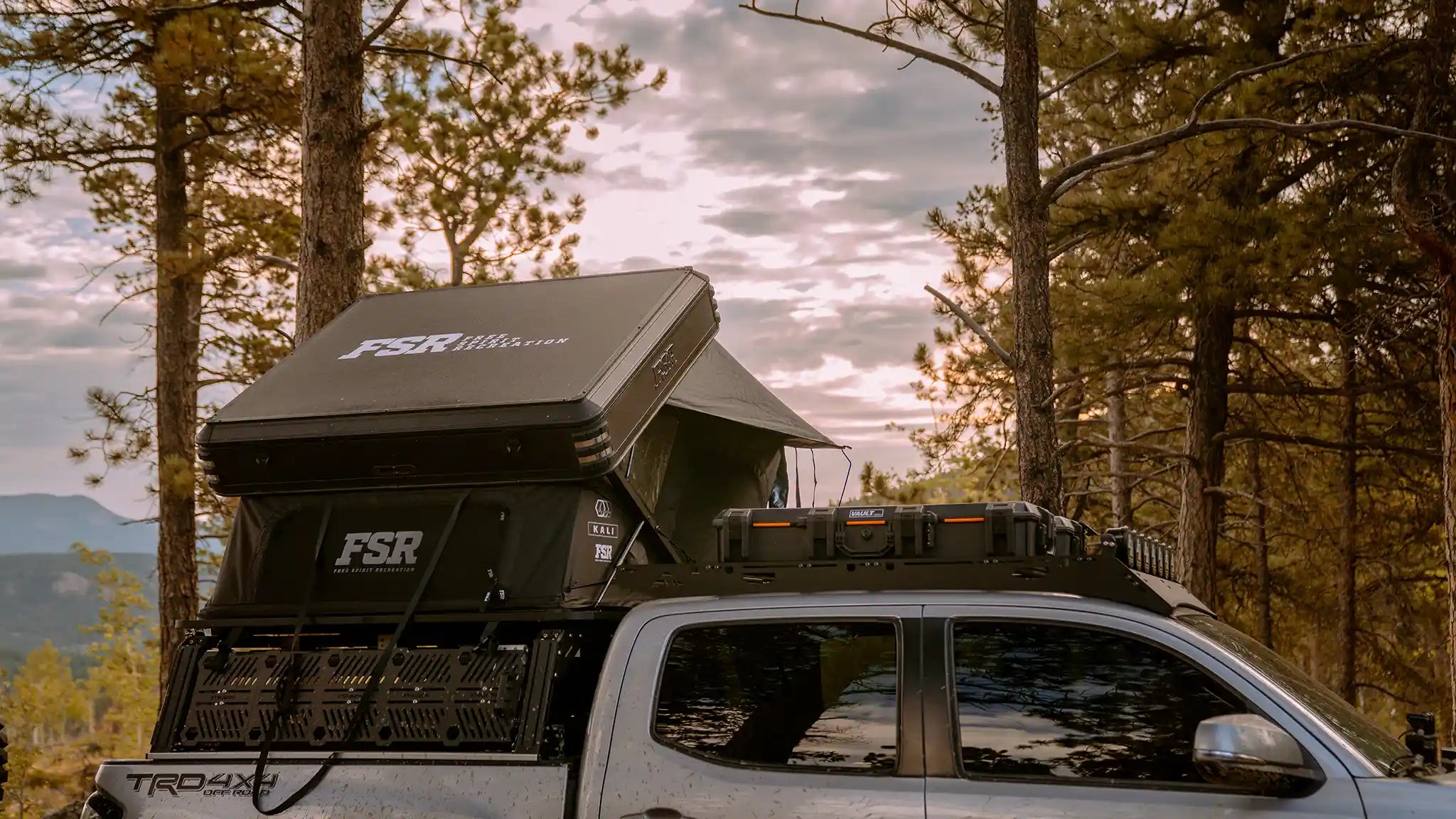 Toyota Tacoma with an overland bed rack mounted FSR kali roof tent setup in a forested campsite with a clouded sky in the background