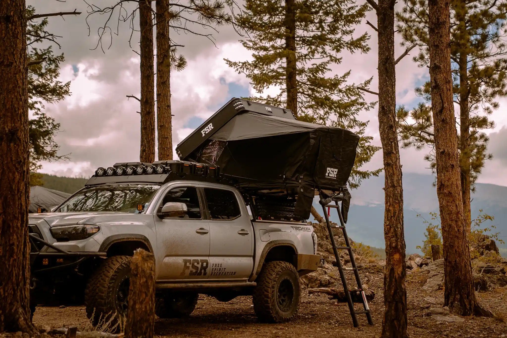 FSR Kali Rooftop Tent from Free Spirit Recreation installed on a Toyota Tacoma bed rack and setup in a wooded campsite with mountains in the background