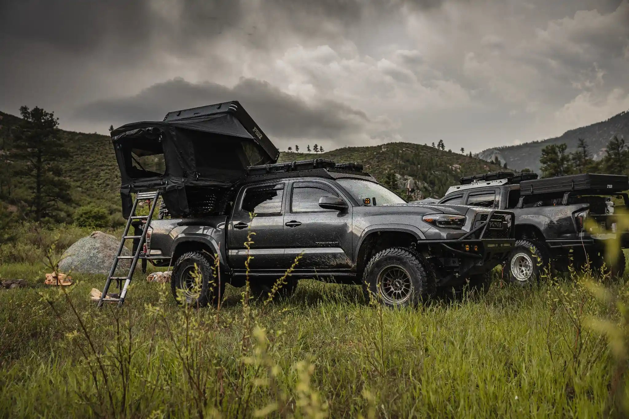 Two Toyota overland trucks with FSR Kali Roof Top Tents setup in a grassy field with mountains in the background