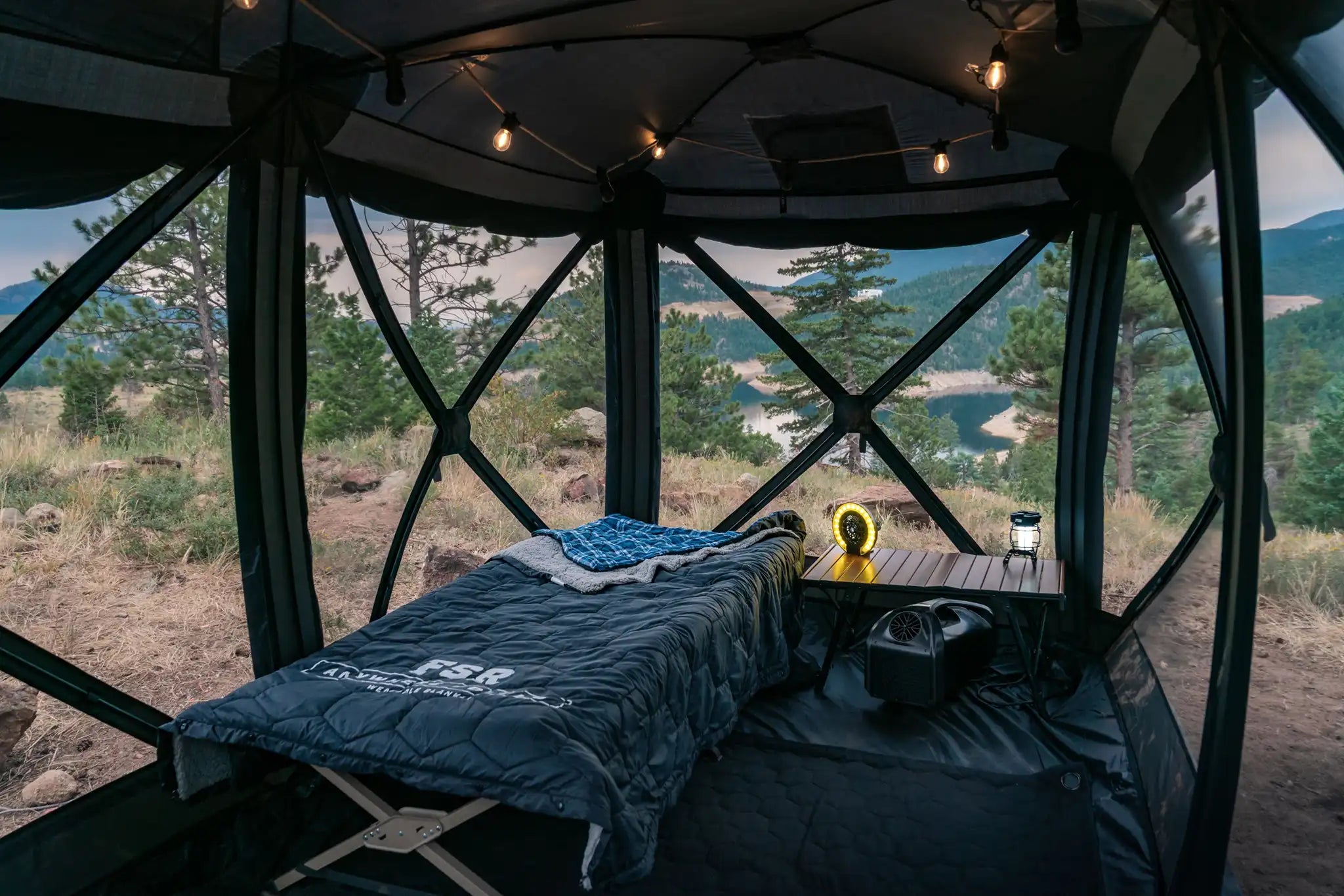 A Free Spirit Recreation Hub Tent setup in a mountainous wooded campsite with windows open and an FSR sleeping bag on a camping cot surrounded by several FSR camping products