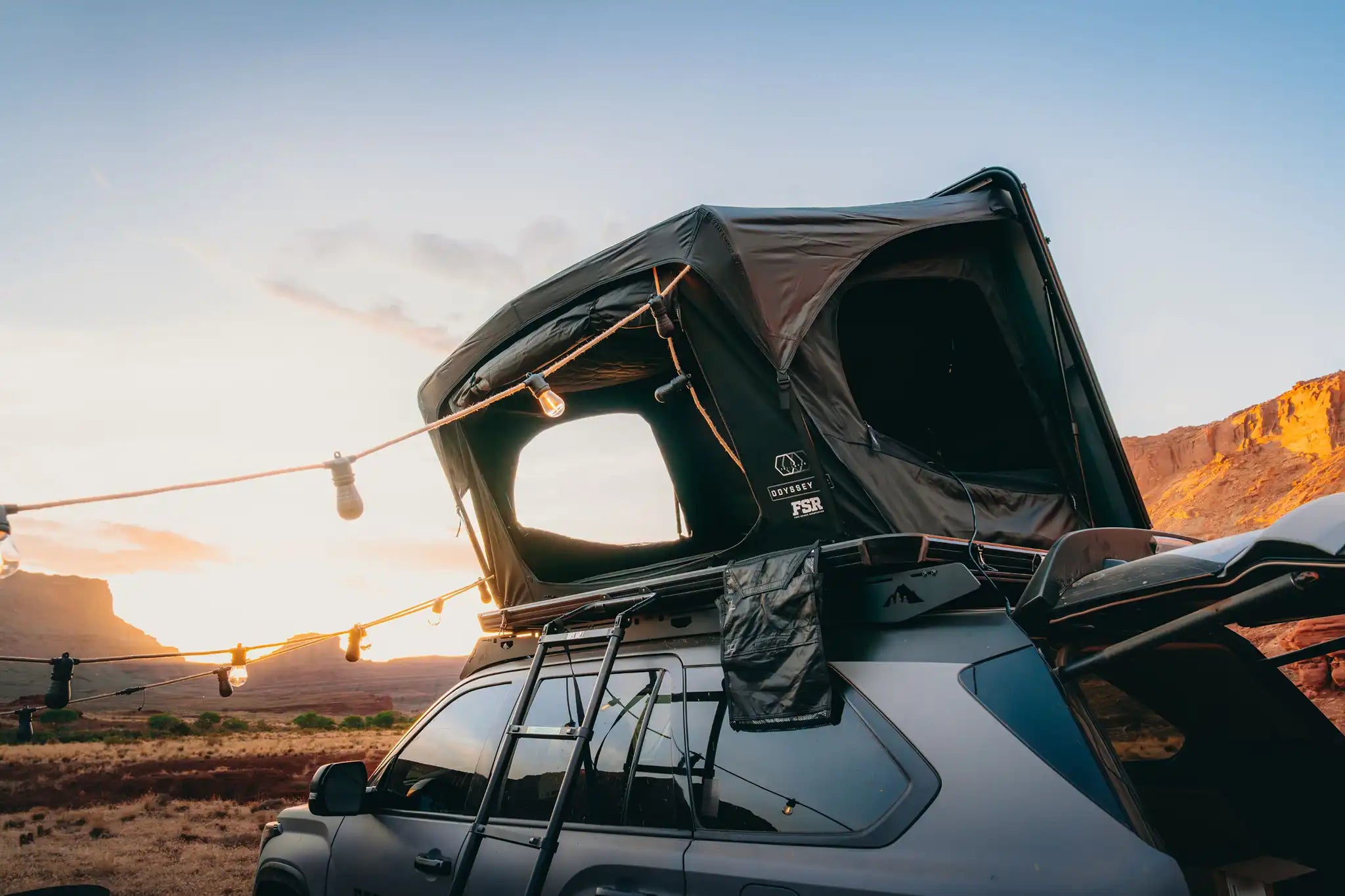 Car with a FSR rooftop tent from Free Spirit Recreation mounted on the roof rack setup for camping in a desert landscape during sunset.