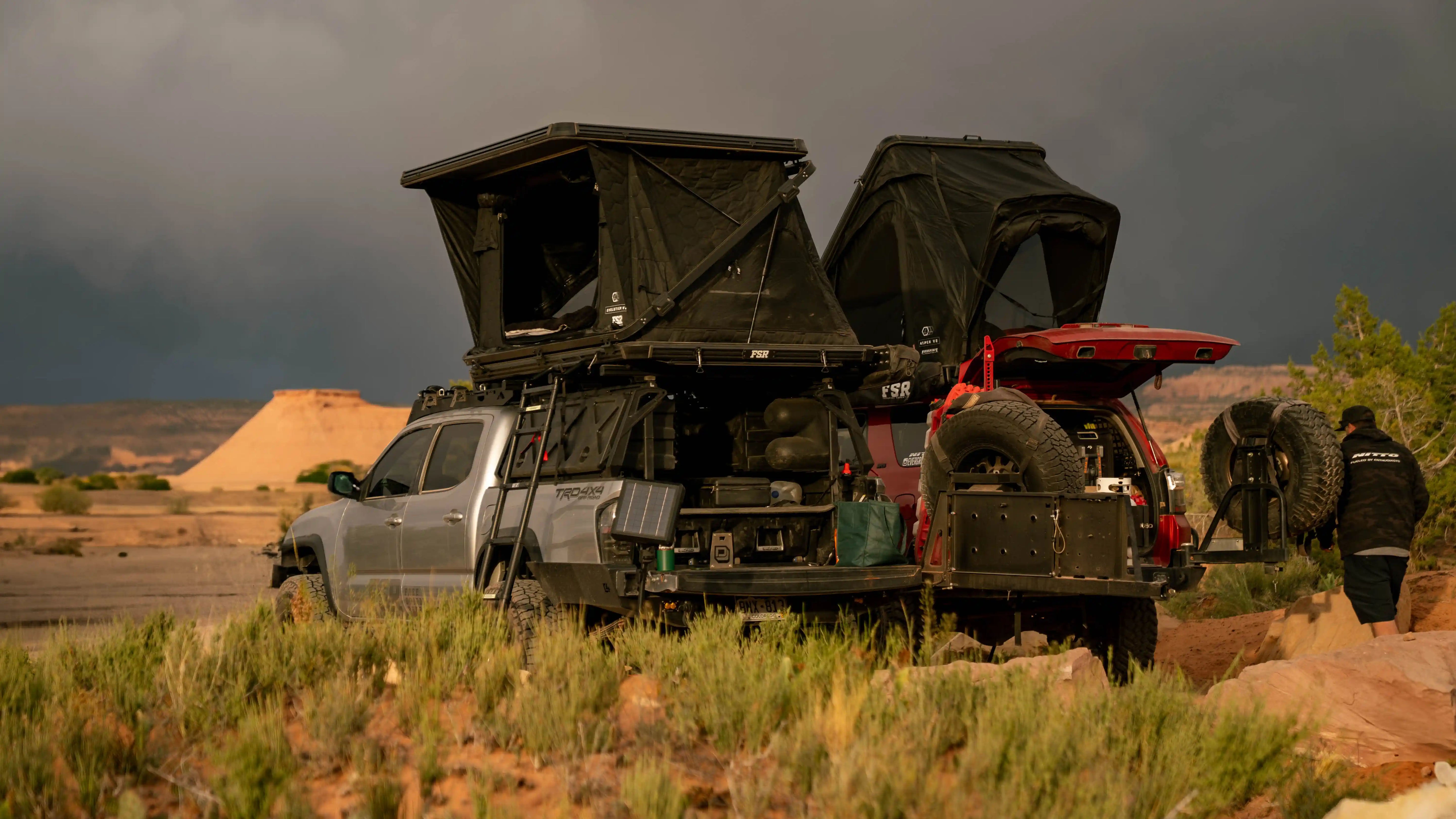 Two vehicles with FSR rooftop tents mounted to roof racks setup in a desert landscape under a stormy sky.
