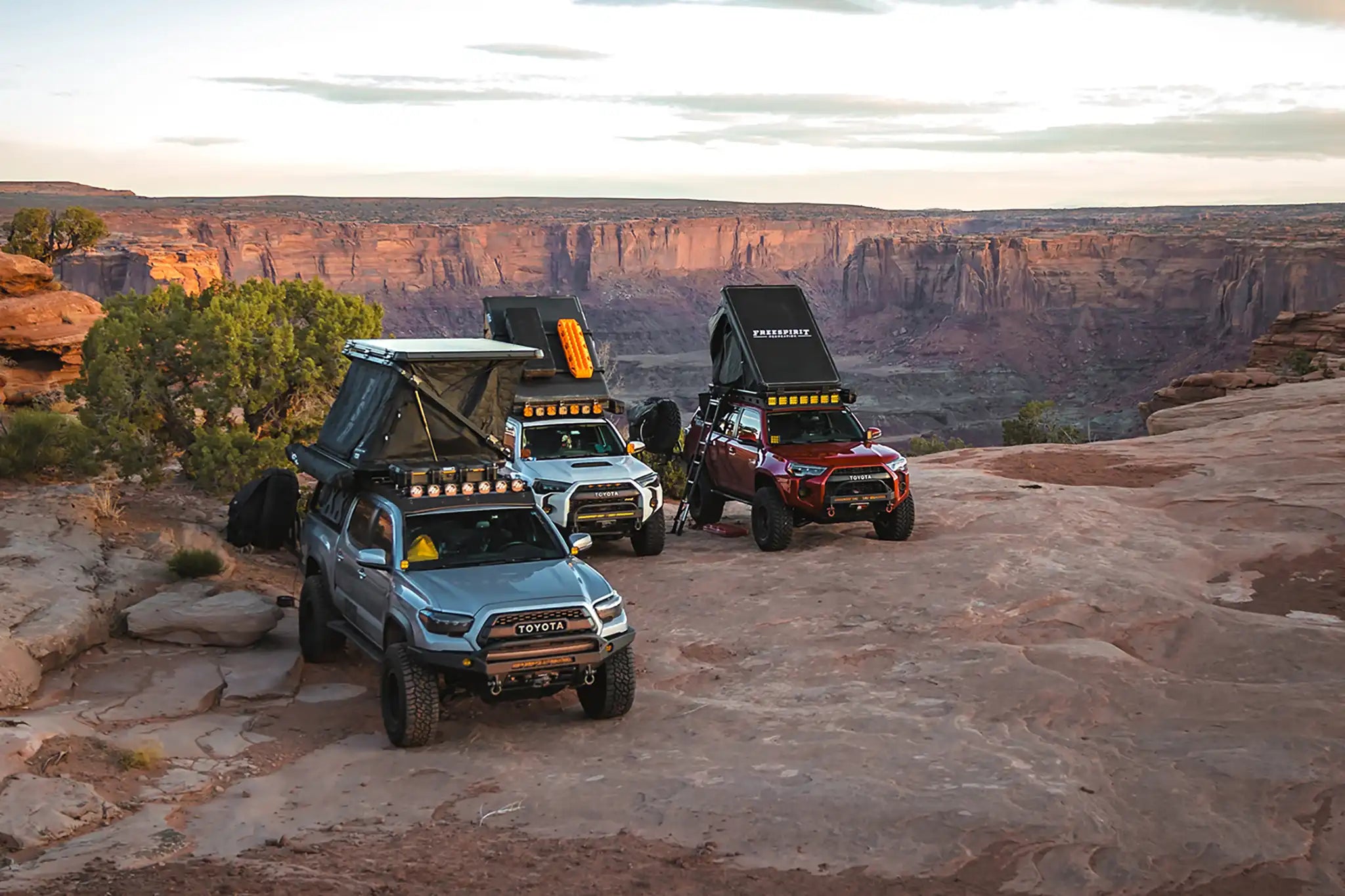 A Toyota Tacoma pickup and two Toyota 4Runner suvs with various FSR rooftop tents from Free Spirit Recreation parked and setup for camping on a rocky bluff overlooking a canyon.