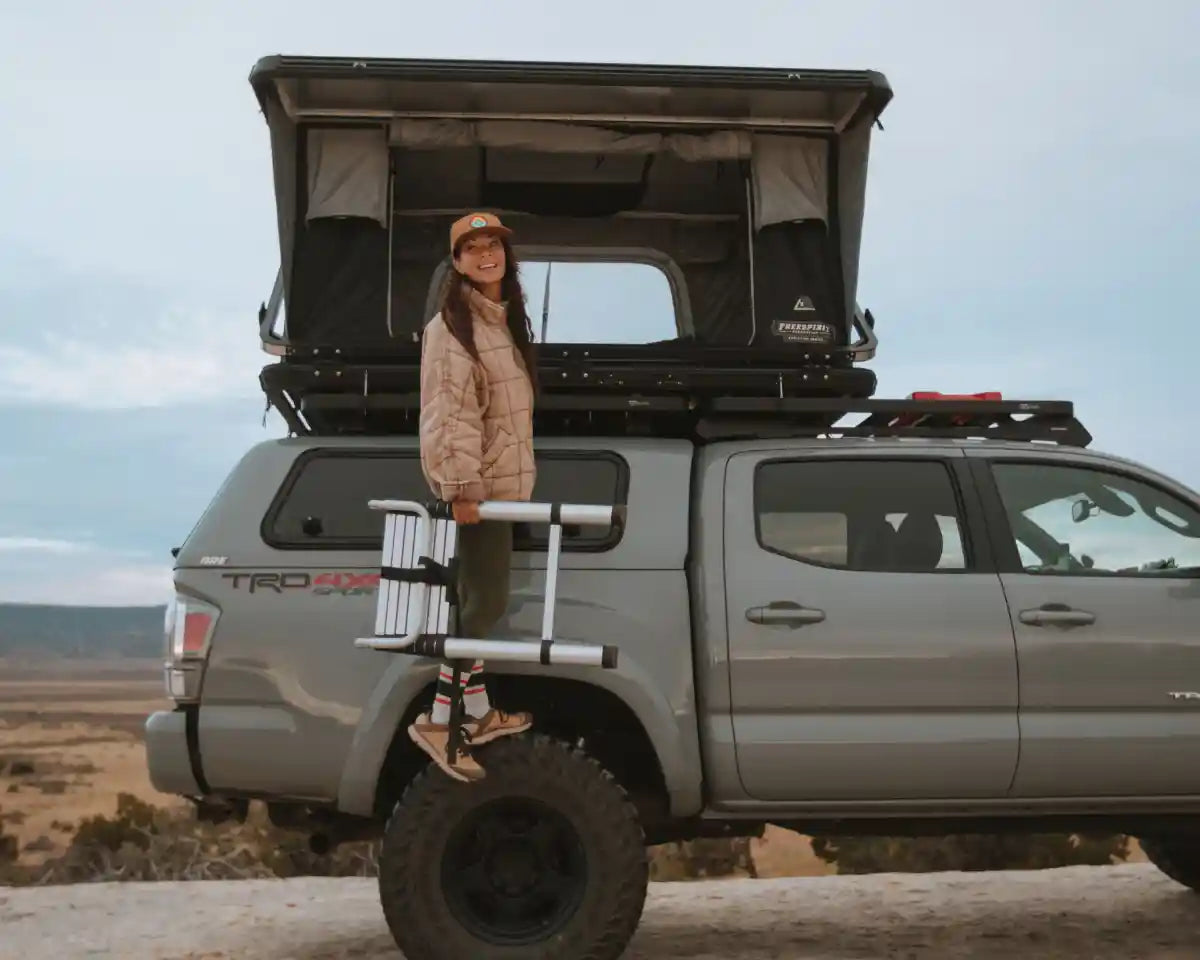 Person standing next to a Toyota Tacoma with an open FSR roof top tent installed on the roof rack, camping in a desert landscape.
