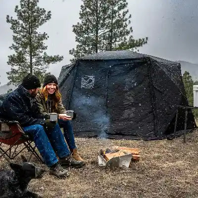 Two people sitting by a campfire in front of an FSR Hub pop-up camping tent with trees in the background