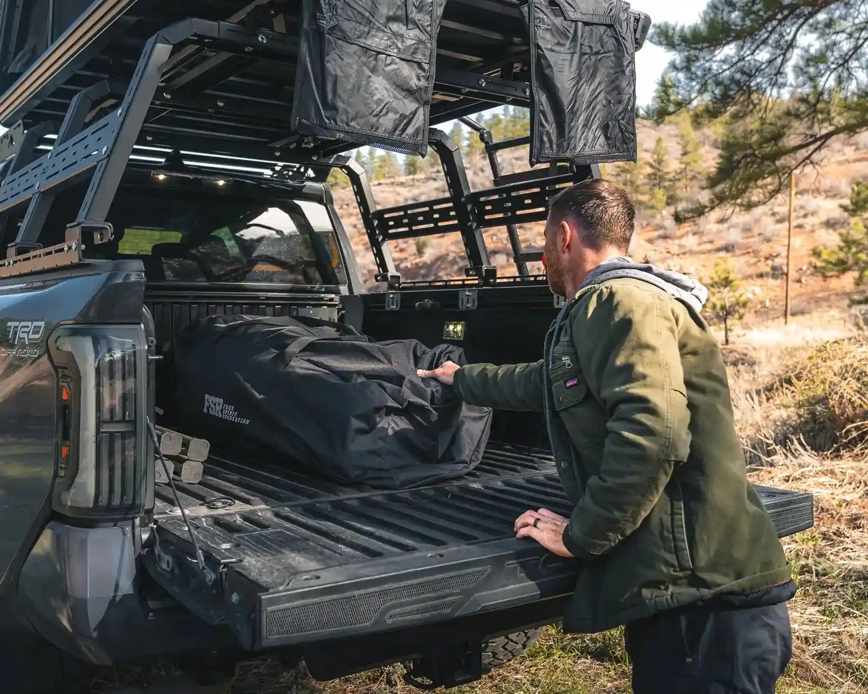 Man removing FSR roof top tent accessories from the back of a truck bed while setting up camp in a sparsly wooded area