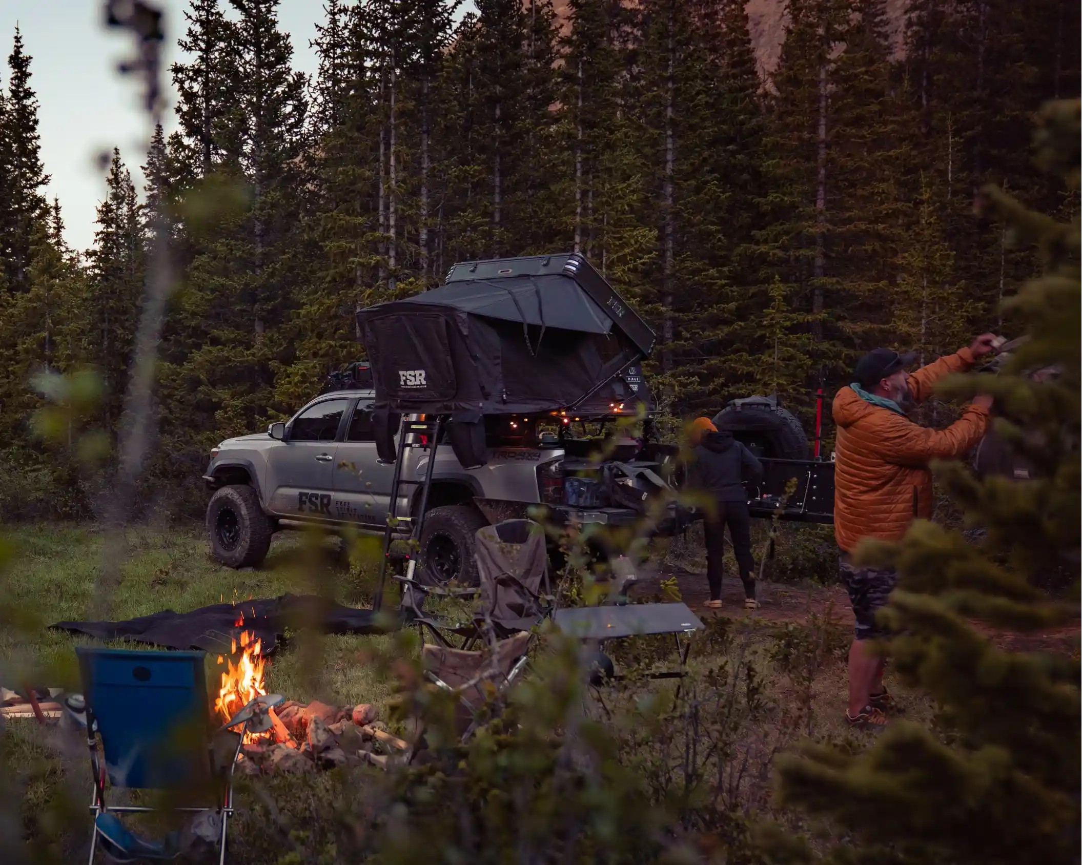 People camping outdoors with a truck and FSR Kali roof tent from Freespirit Recreation in a forest setting with a campfire visible in the foreground