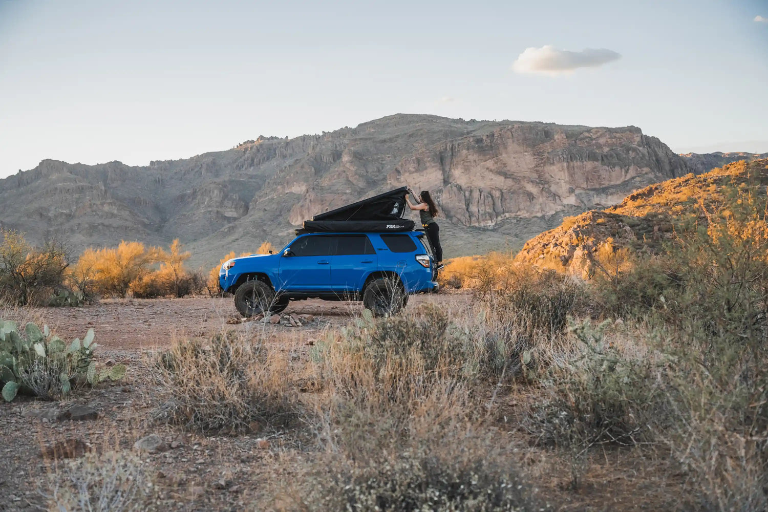Person opening a clam shell roof top tent from Free Spirit Recreation mounted on top of thier blue Toyota 4Runner in a desert campsite at sunset with mountains in the background