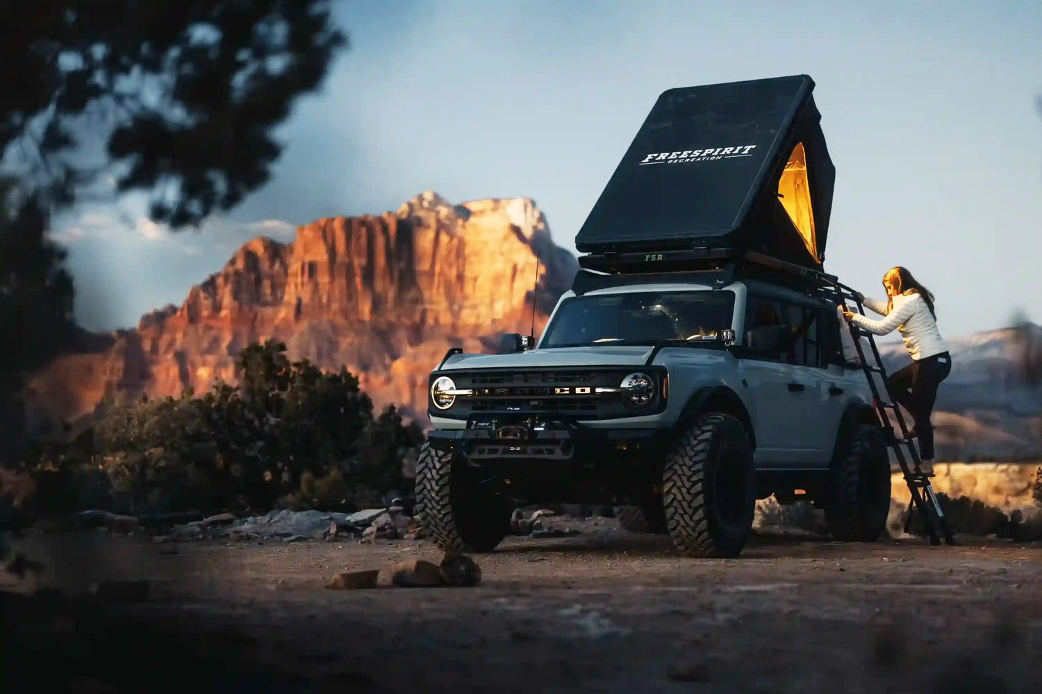 Person climbing into thier Ford Bronco roof tent from Free Spirit Recreation setup in a scenic desert campsite at dusk with sun setting on a red rock formation in the background