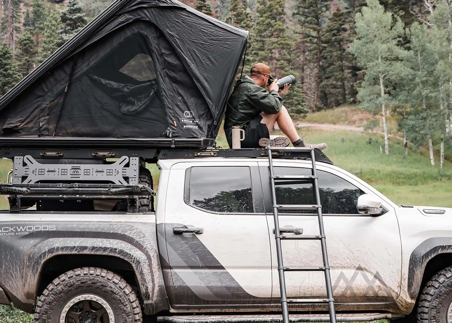Man sitting on top of a Short bed Toyota Tacoma pickup with an FSR Rooftop Tent from Free Spirit Recreation mounted above the cab on a full-heght bed rack and setup while camping in a wooded area