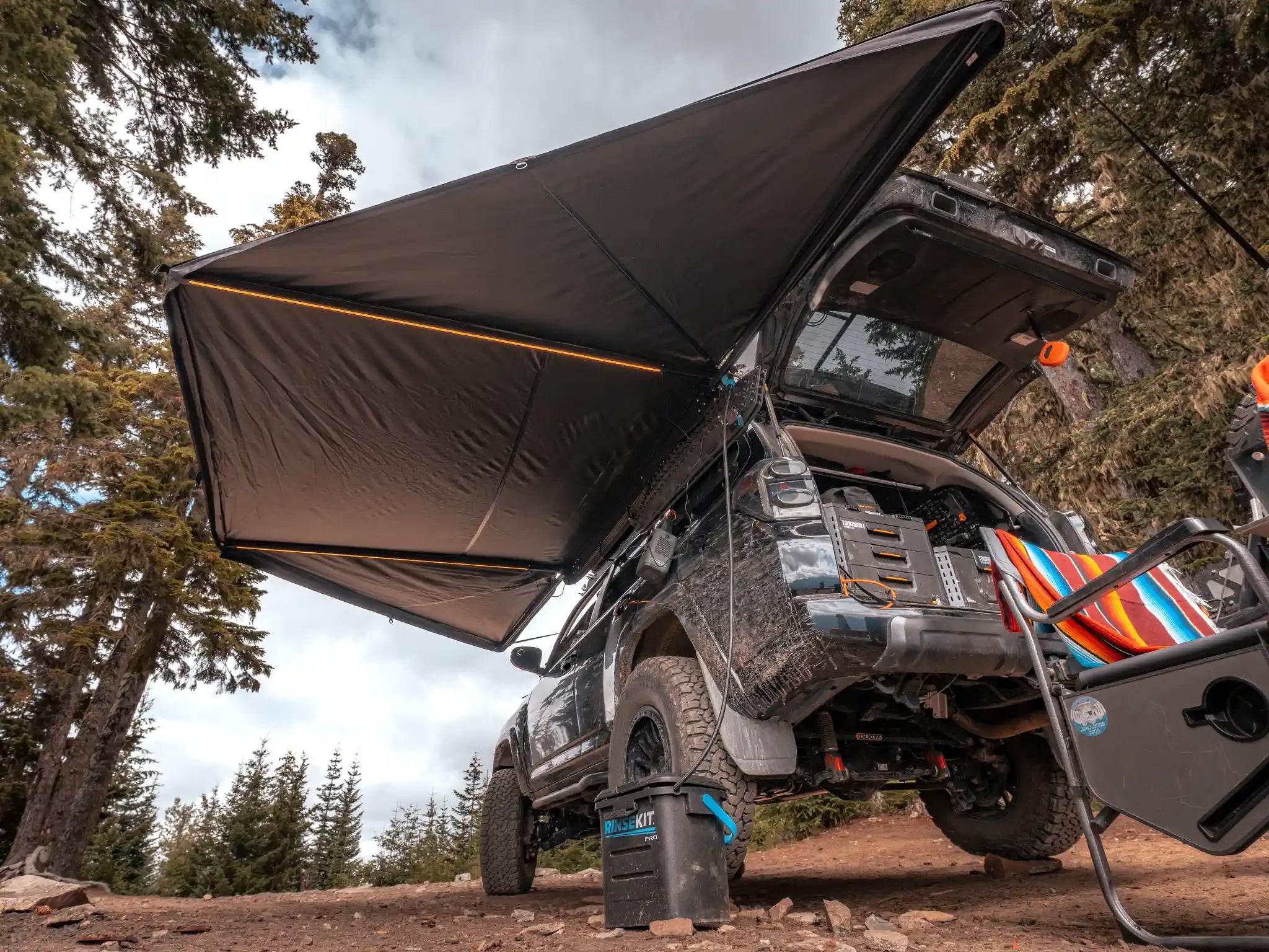 Black Toyota 4Runner with a FSR 180 degree Awning V2 from Free Spirit Recreation setup camping in a wooded area with the car awning deployed and built-in LED lights illuminated.