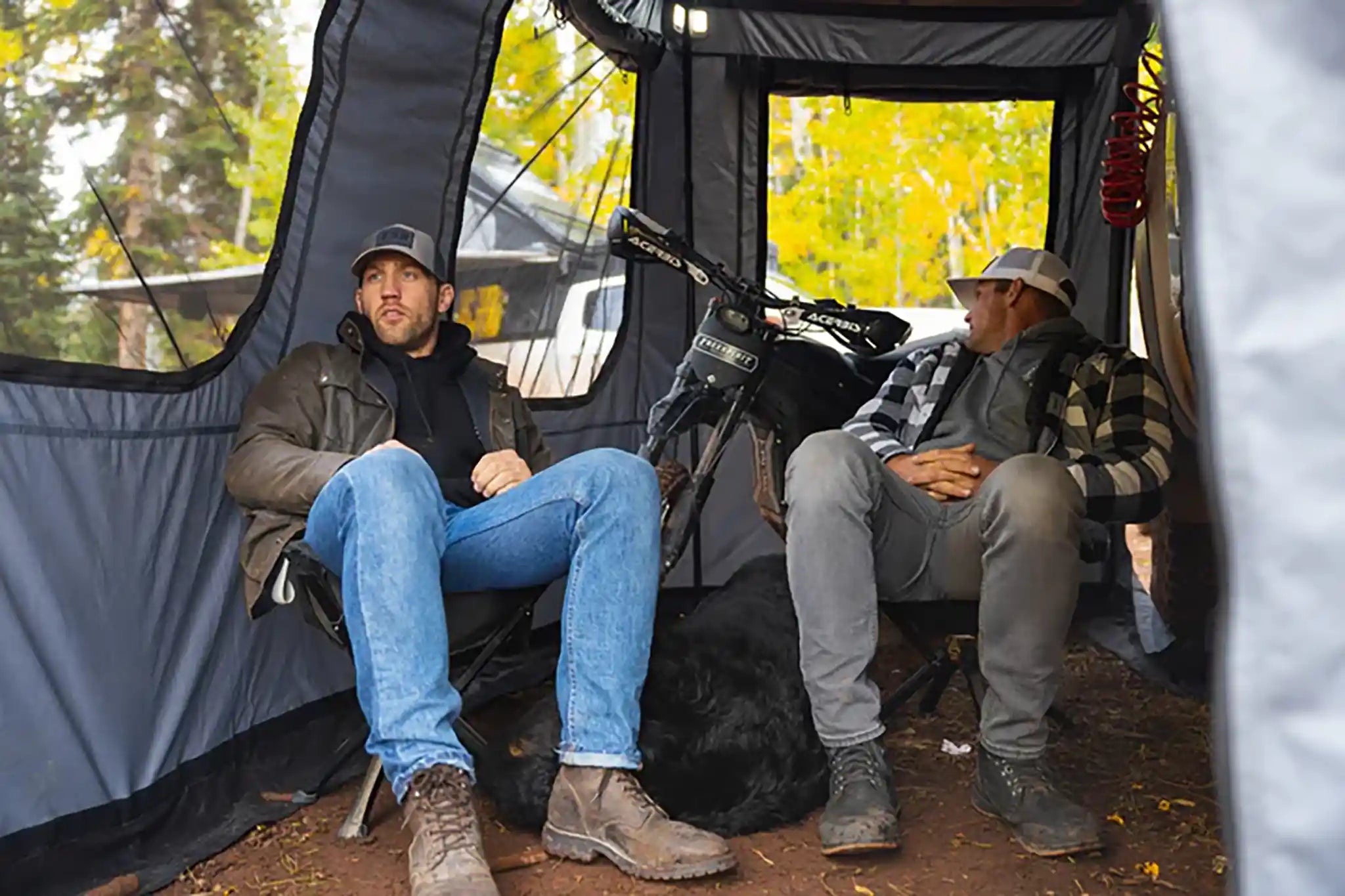 Two men sitting inside an FSR Hub pop-up camping tent with a dog and a dirt bike, autumn foliage visible through the tent's open windows.