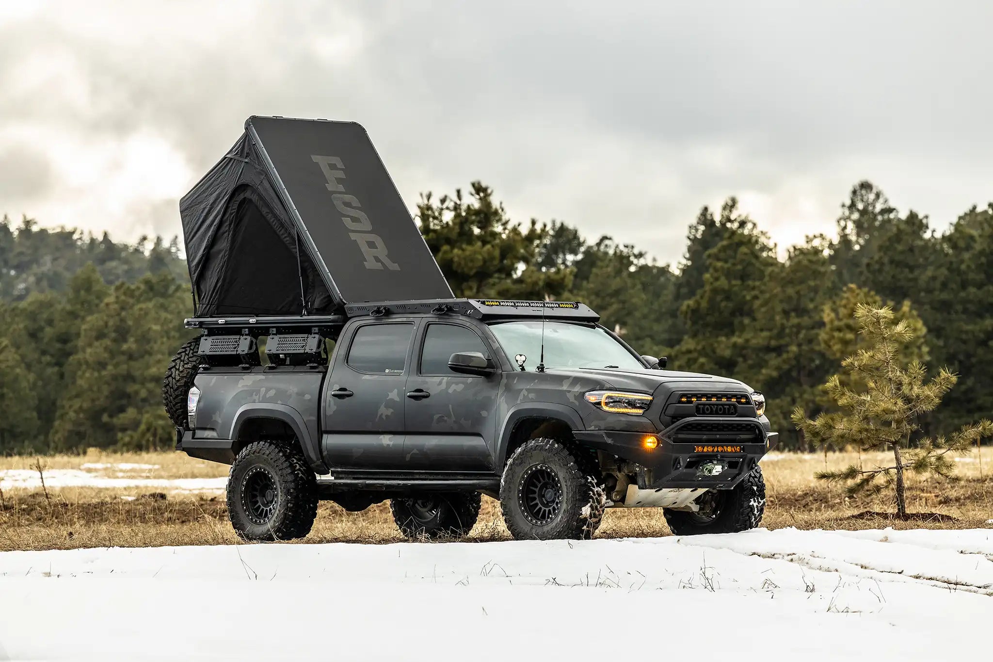 Person camping in an FSR Aspen Lite XL Rooftop Tent installed on a overland built Toyota Tacoma in a snow covered field with forest in the background