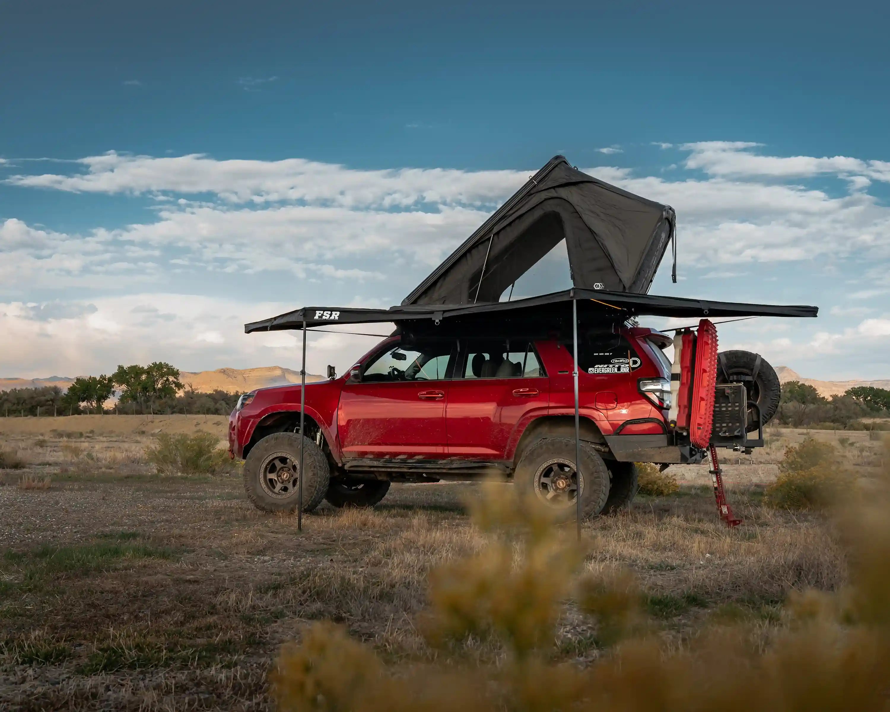 Red Toyota 4Runner with a Freespirit Recreation rooftop tent and car awning setup for camping in an open field under a blue sky with a mountain range visible in the background.