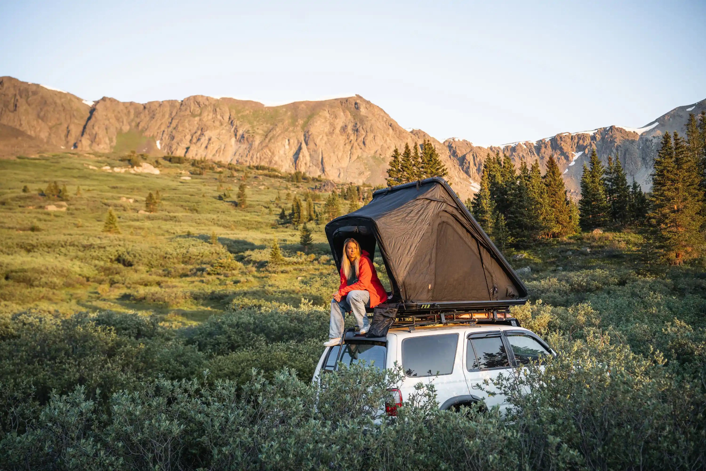 Person sitting on an FSR roof tent  mounted on top of a car camping in a mountainous landscape