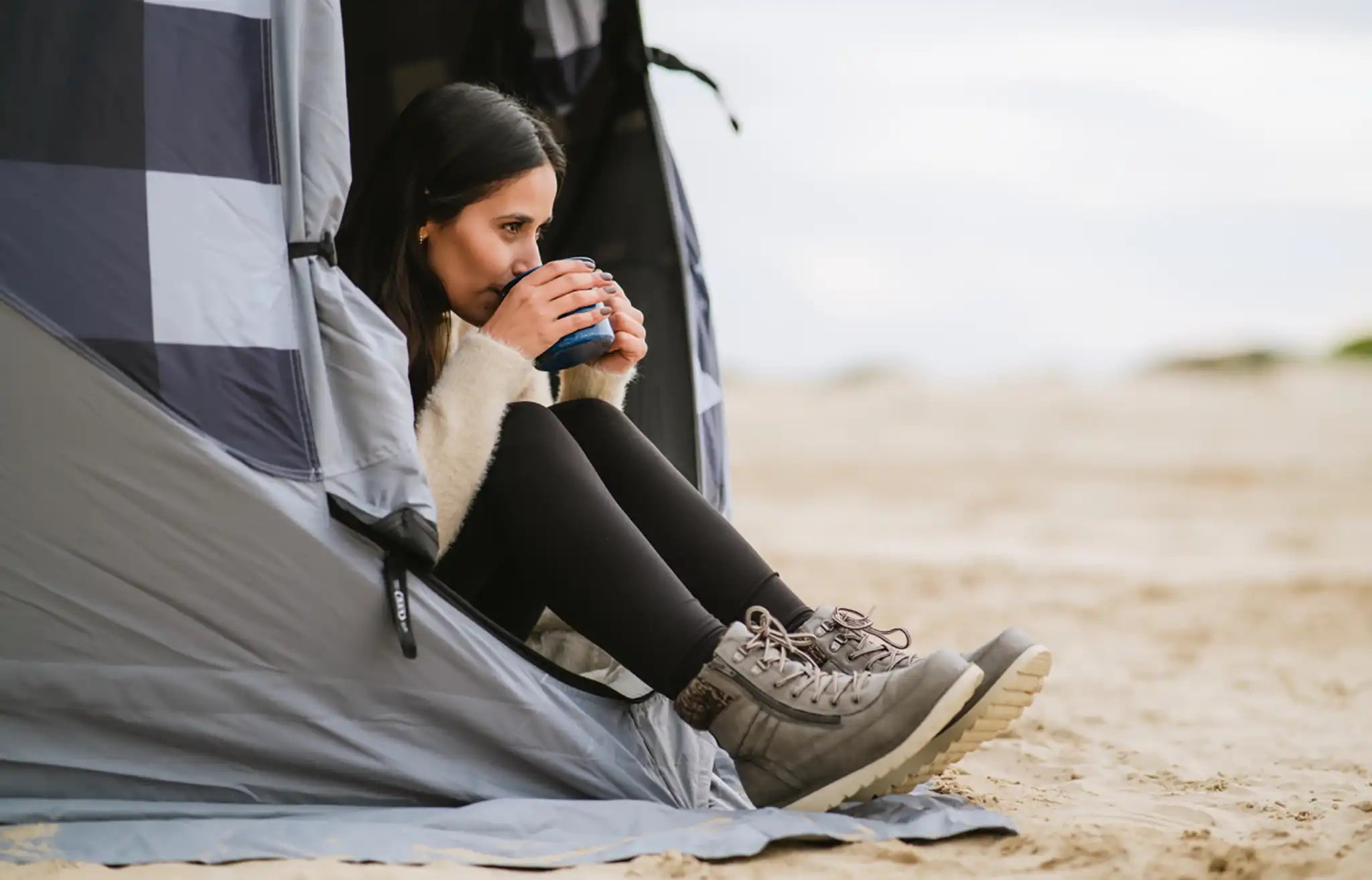 Woman sitting in the entry door of a FSR Hub pop-up camping tent drinking coffee from a blue mug on a beach.
