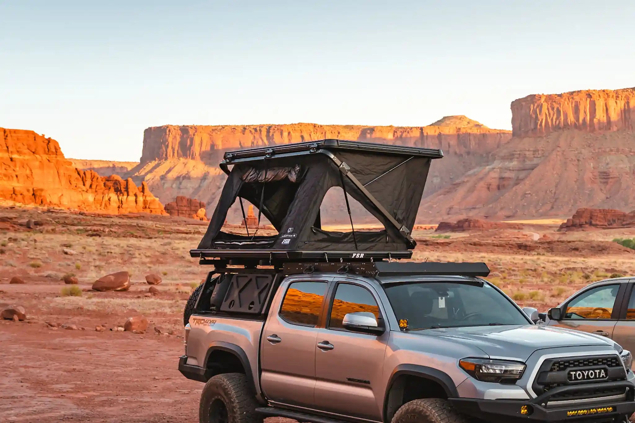 FSR Evolution V2 Rooftop Tent from Free Spirit Recreation installed and setup on a gray Toyota Tacoma bed rack in a desert campsite surrounded by red rock formations