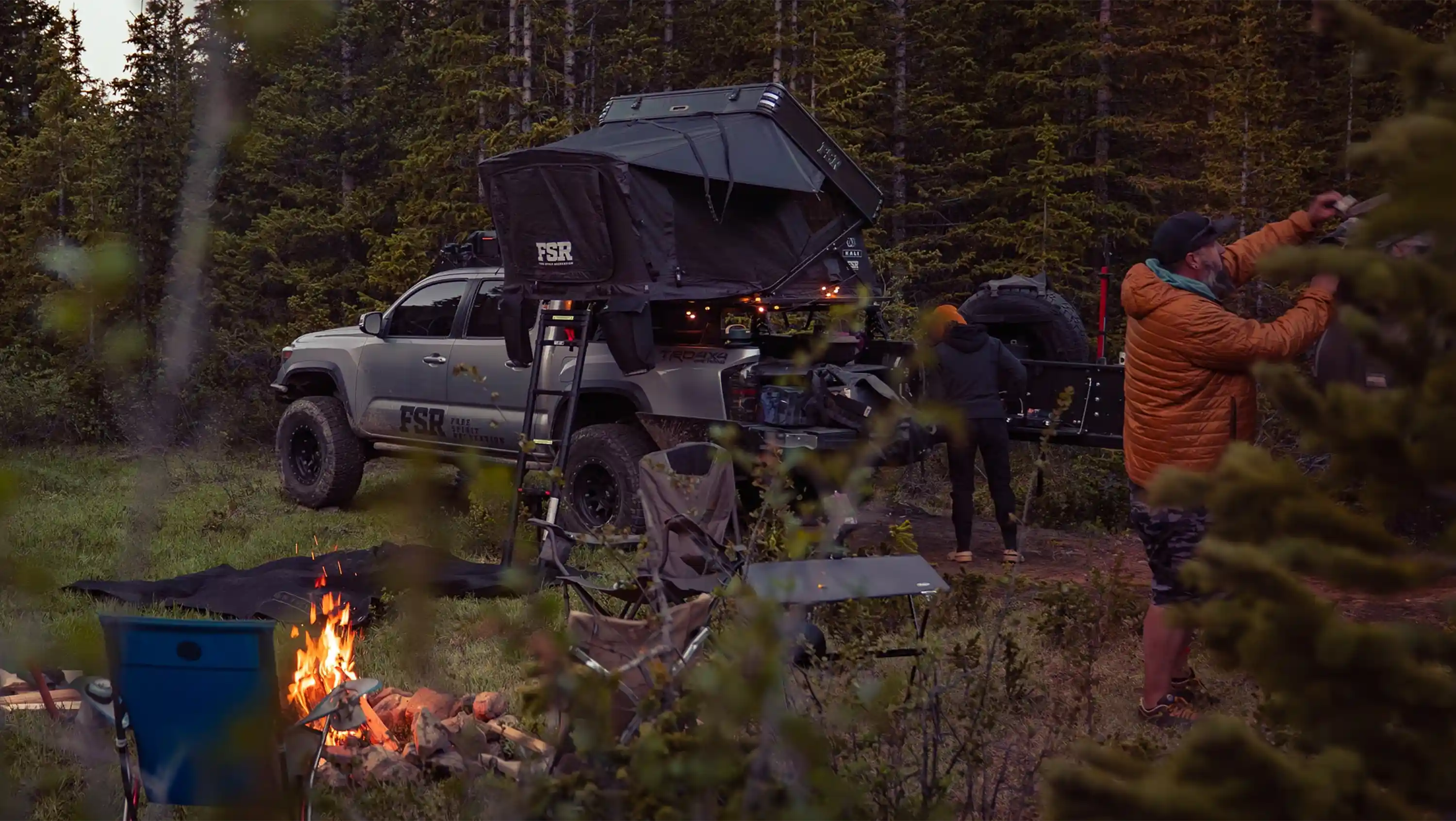 People camping outdoors with a truck equipped with an FSR roof tent in a forest at night.
