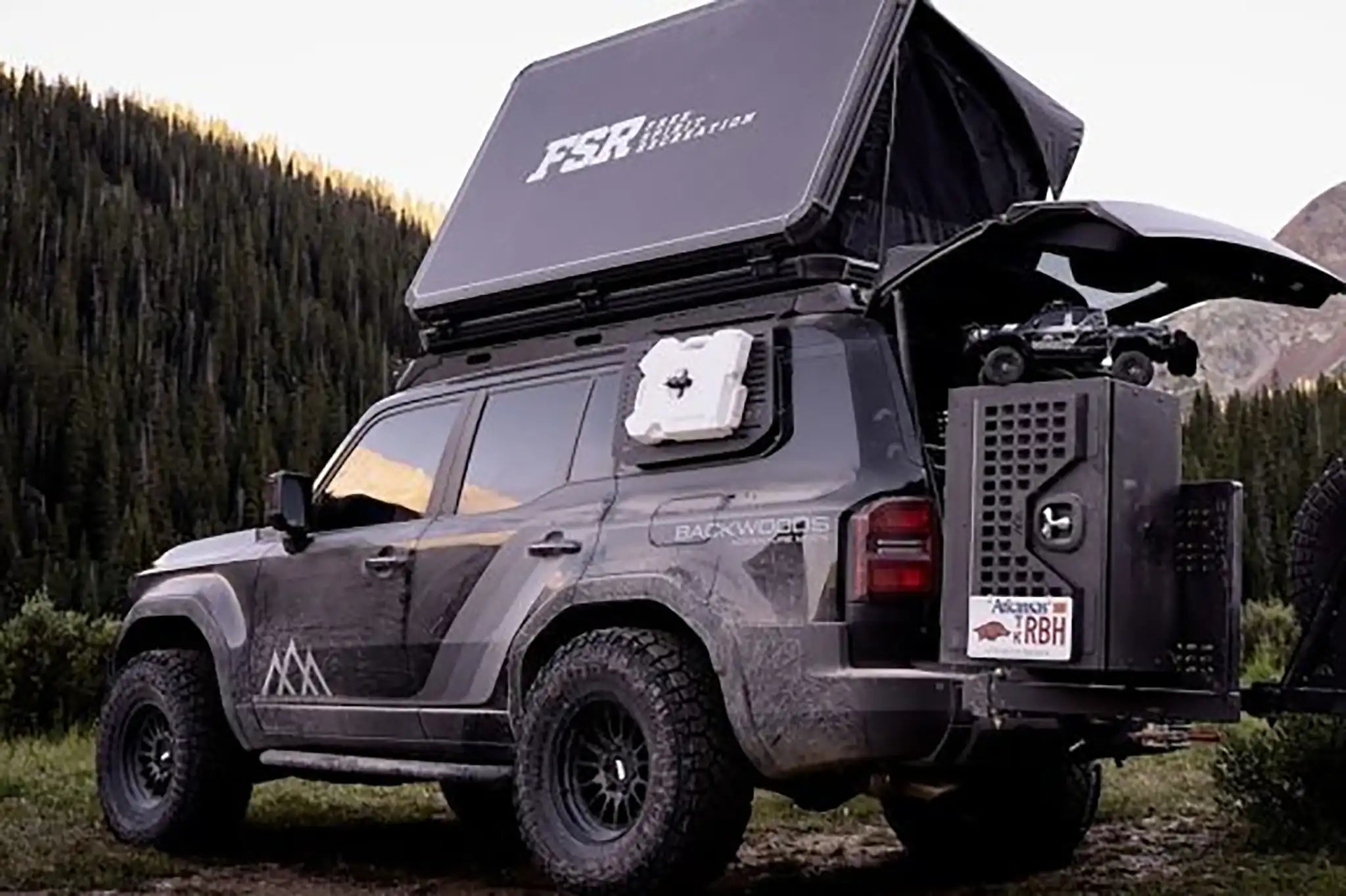 FSR Rooftop Tent from Free Spirit Recreation installed on a Landcruiser 250 with Backwoods Adventure Mods off road and overland accessories parked on a forested campsite with mountains in the background