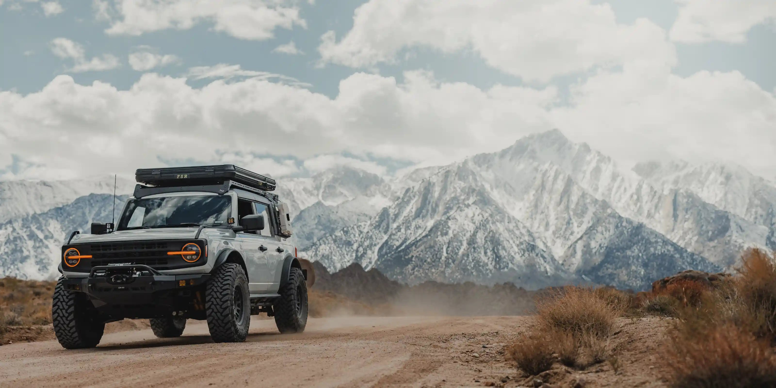 Ford Bronco equipped with an FSR Rooftop Tent from Free Spirit Recreation mounted on the roof rack driving on a dirt road with snow covered mountains in the background