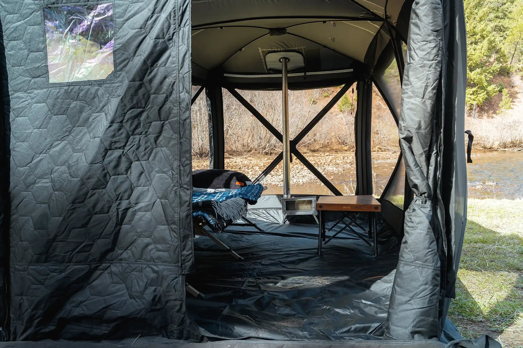 Person camping in the FSR Hub 4 Double Tent, a pop up family camping and glamping tent from Freespirit Recreation with stove installed and roll up walls showing a river in the background 