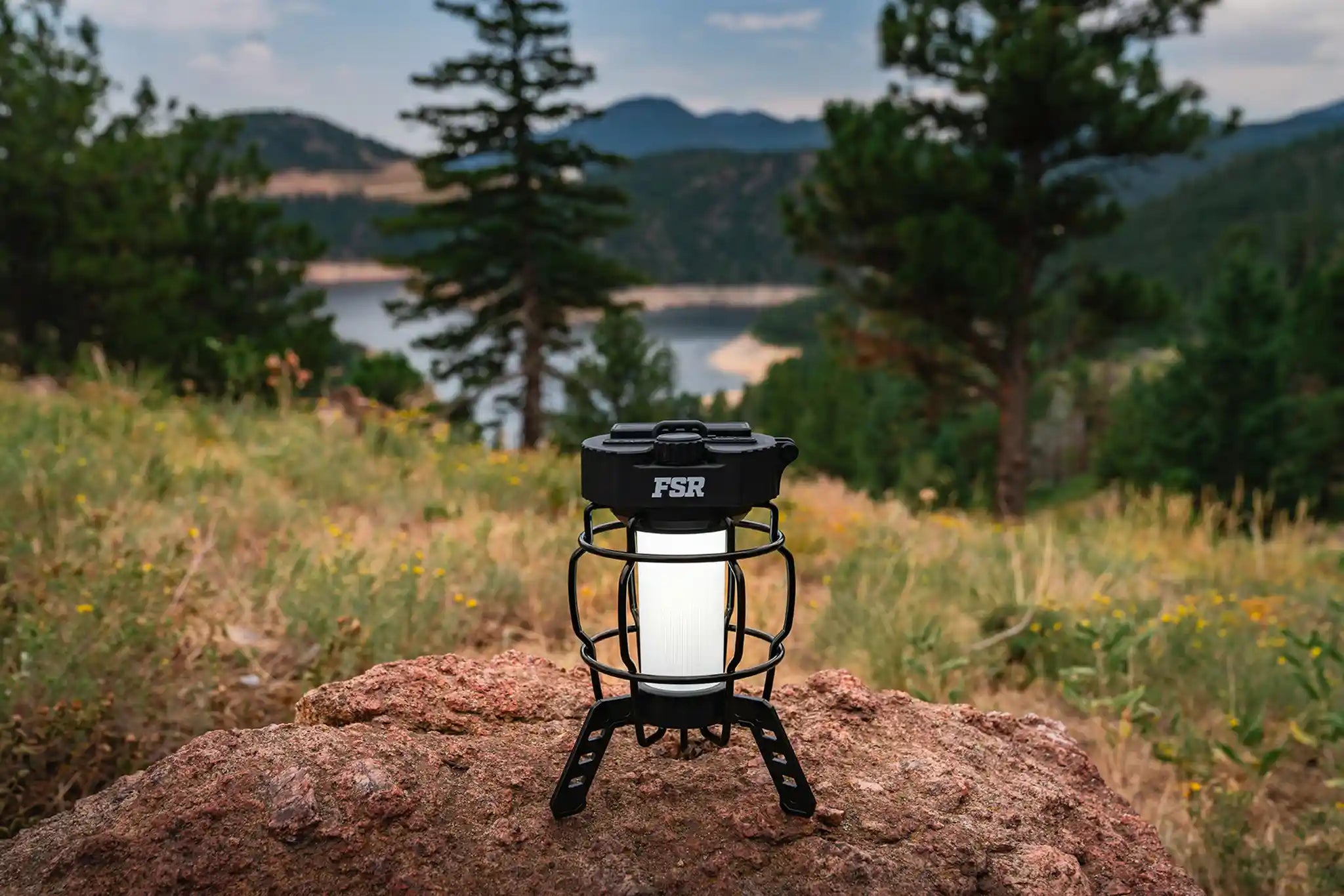 FSR LED Camping Lantern illuminated at a campsite on a rock with a scenic background of pine trees and water