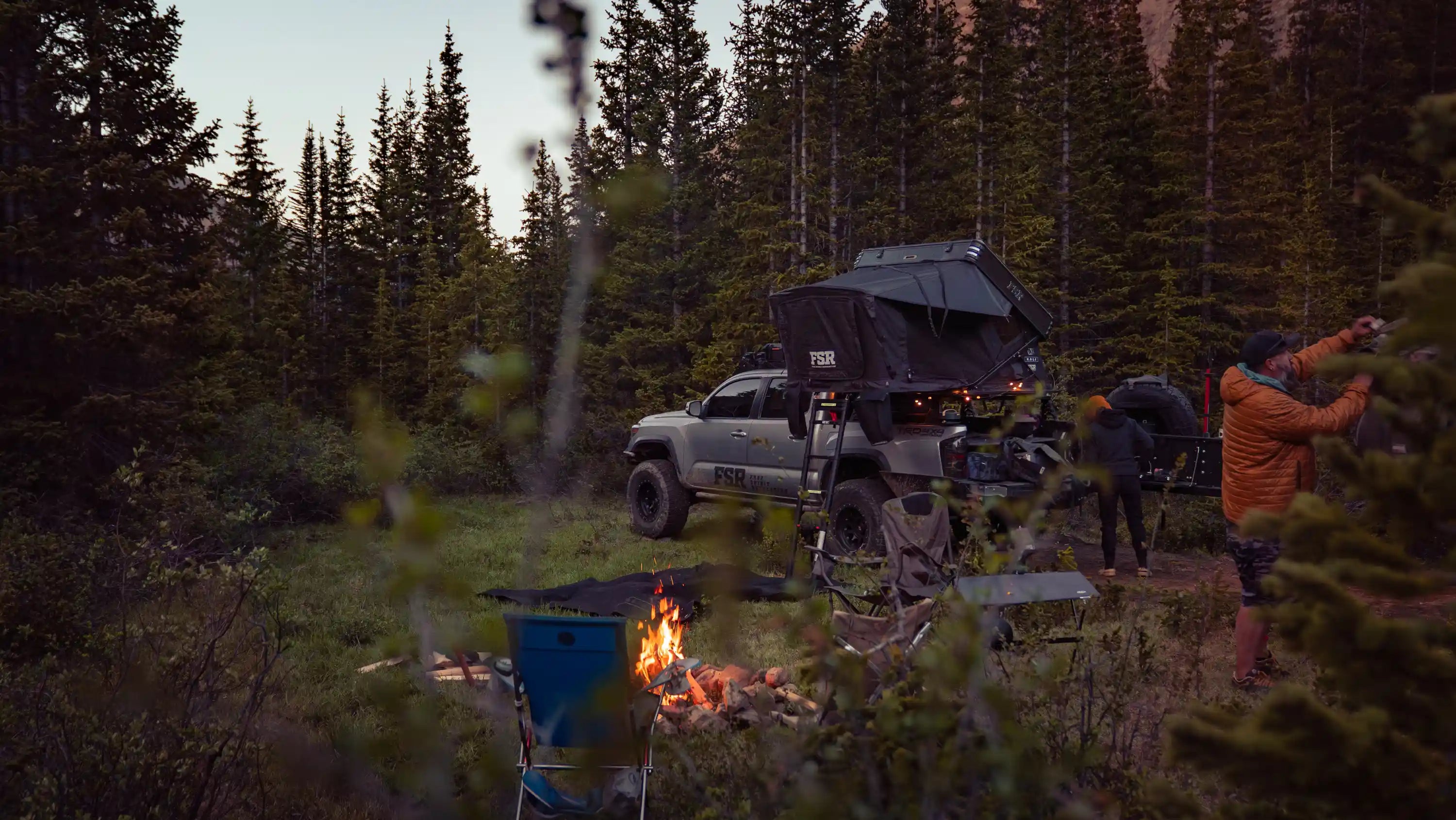 People camping outdoors with a Toyota Tacoma and FSR Rooftop Tent in a forested setting, using portable propane fire pit and campfire cooking equipment from Free Spirit Recreation