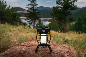 FSR camping lantern setup on a rock in a campsite with a scenic view of trees and a body of water in the background