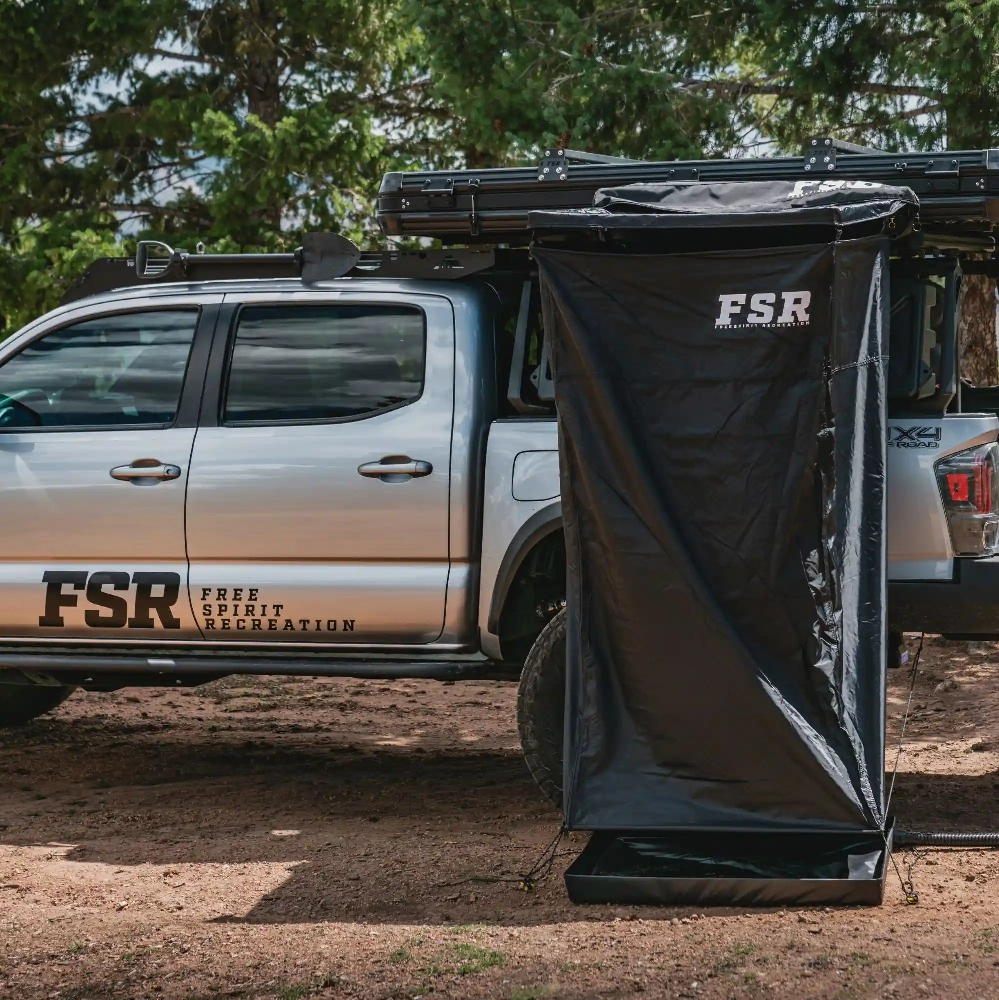 Silver Toyota Tacoma with an FSR shower awning mounted to a FreeSpirit Recreation rooftop tent installed on the truck's bed rack while camped in a forested setting